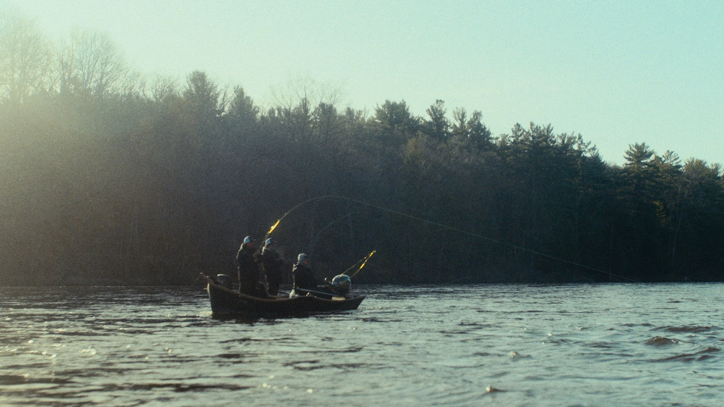 Four people fishing from a boat on a lake near a wooded shoreline during daytime.