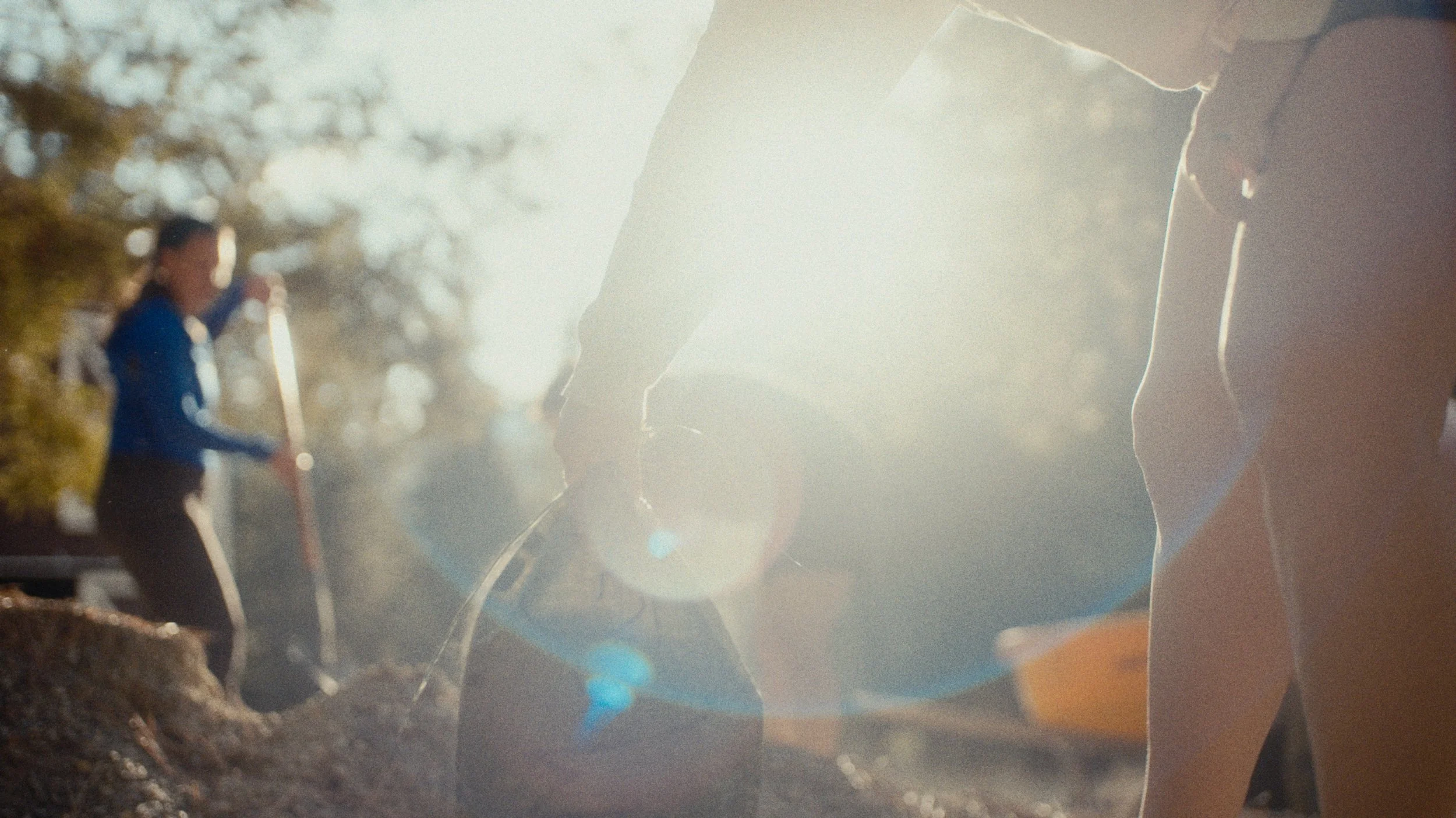 People working outdoors in a sunlit setting, with one person shoveling dirt or soil, and other individuals engaged in activity nearby. The scene appears to be in the late afternoon with sunlight creating a bright, warm glow.