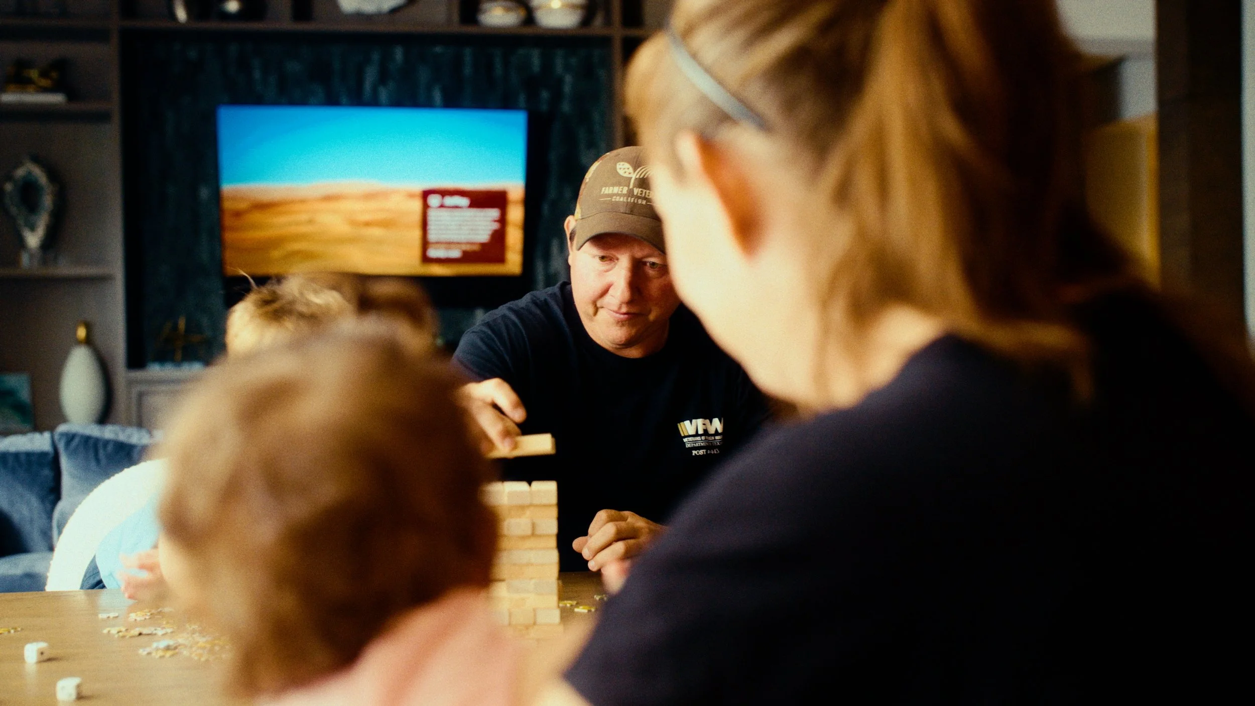 People playing a game of Jenga at a table in a living room, with a TV displaying a landscape in the background.