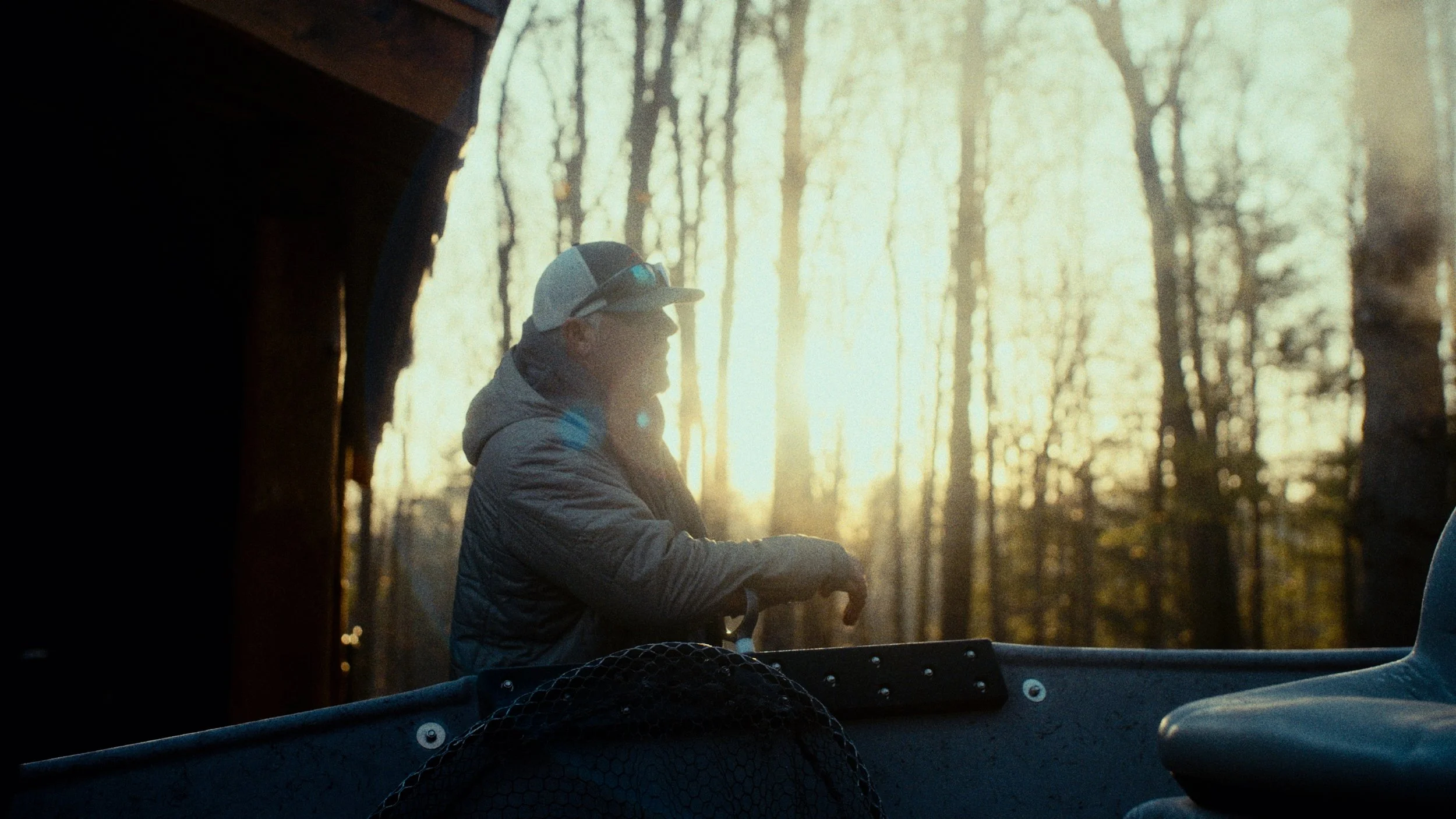 A person sitting in a boat at sunset, wearing a cap and jacket, surrounded by a wooded area with bare trees.