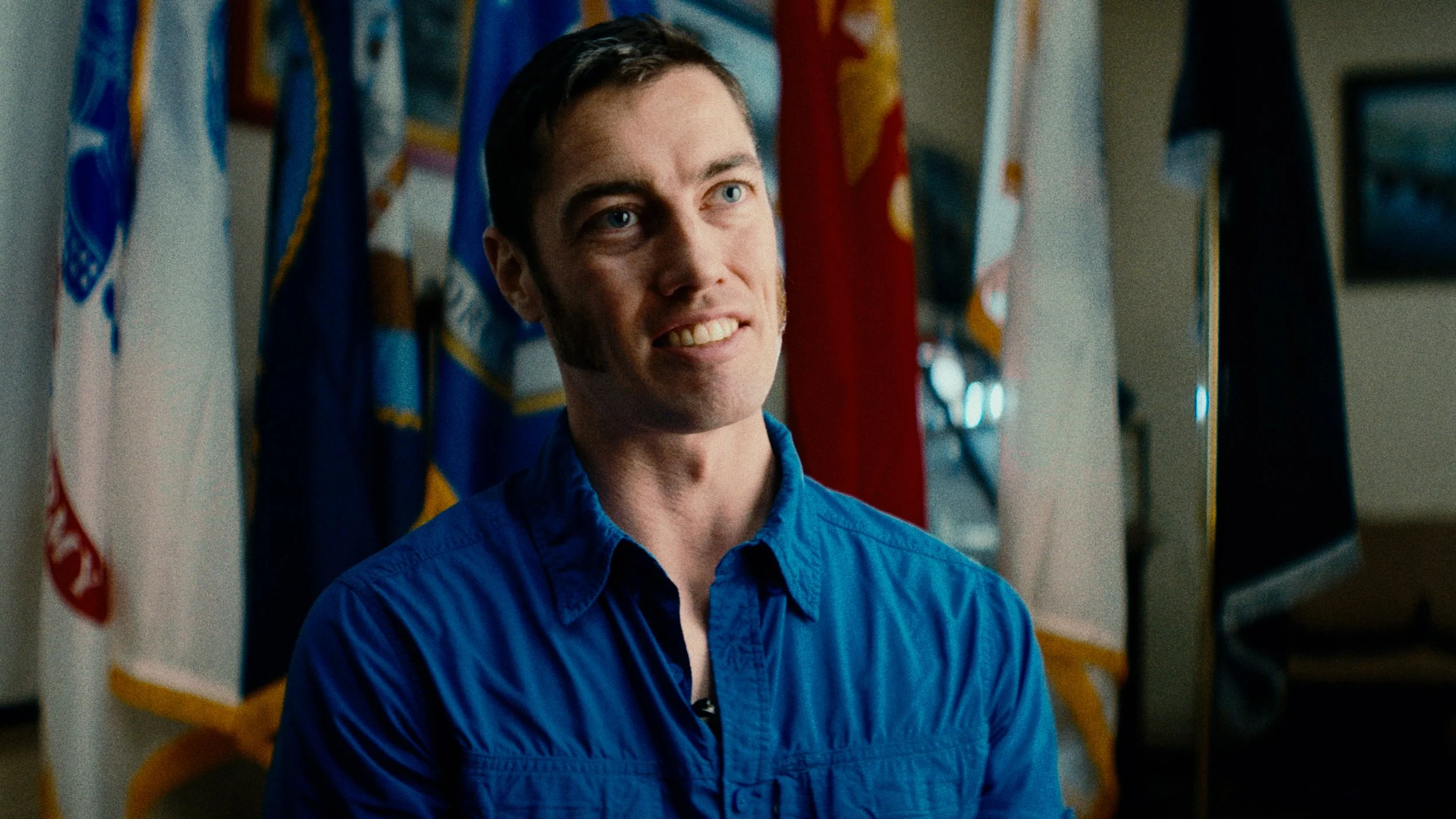 A man with short dark hair and blue eyes, smiling with teeth showing, standing in front of flags with yellow, red, white, and blue colors.