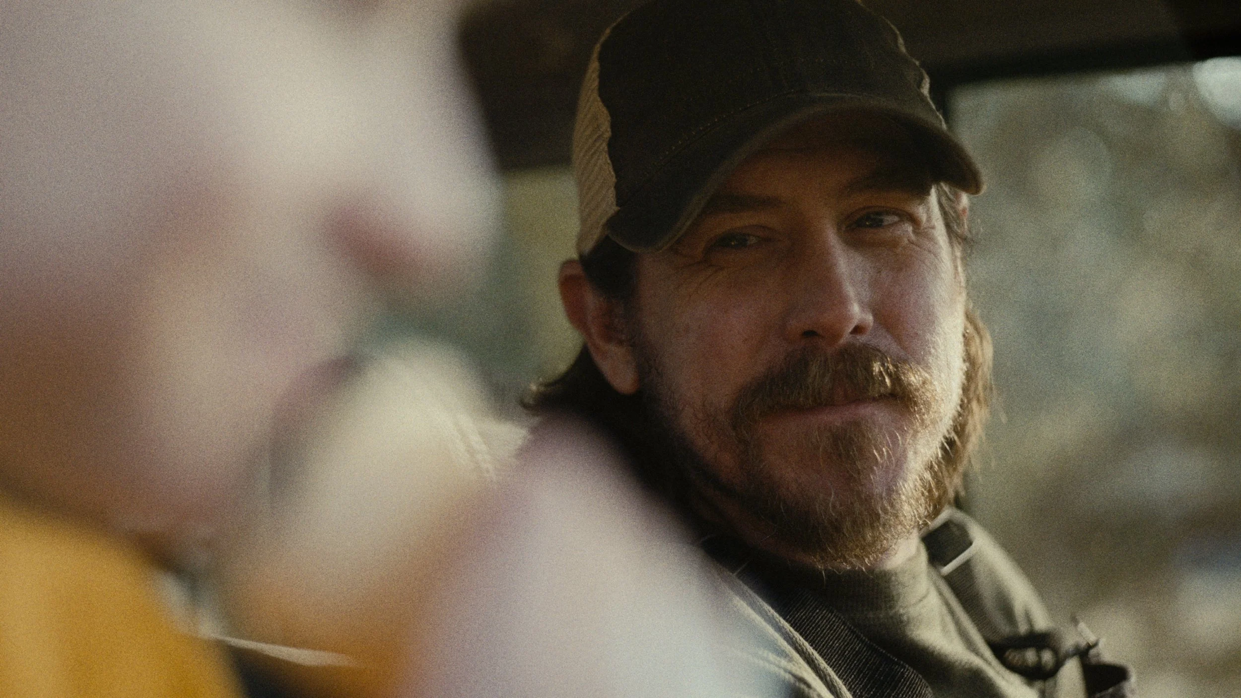 A man with a beard and mustache wearing a baseball cap, looking out of a vehicle window with a slight smile.