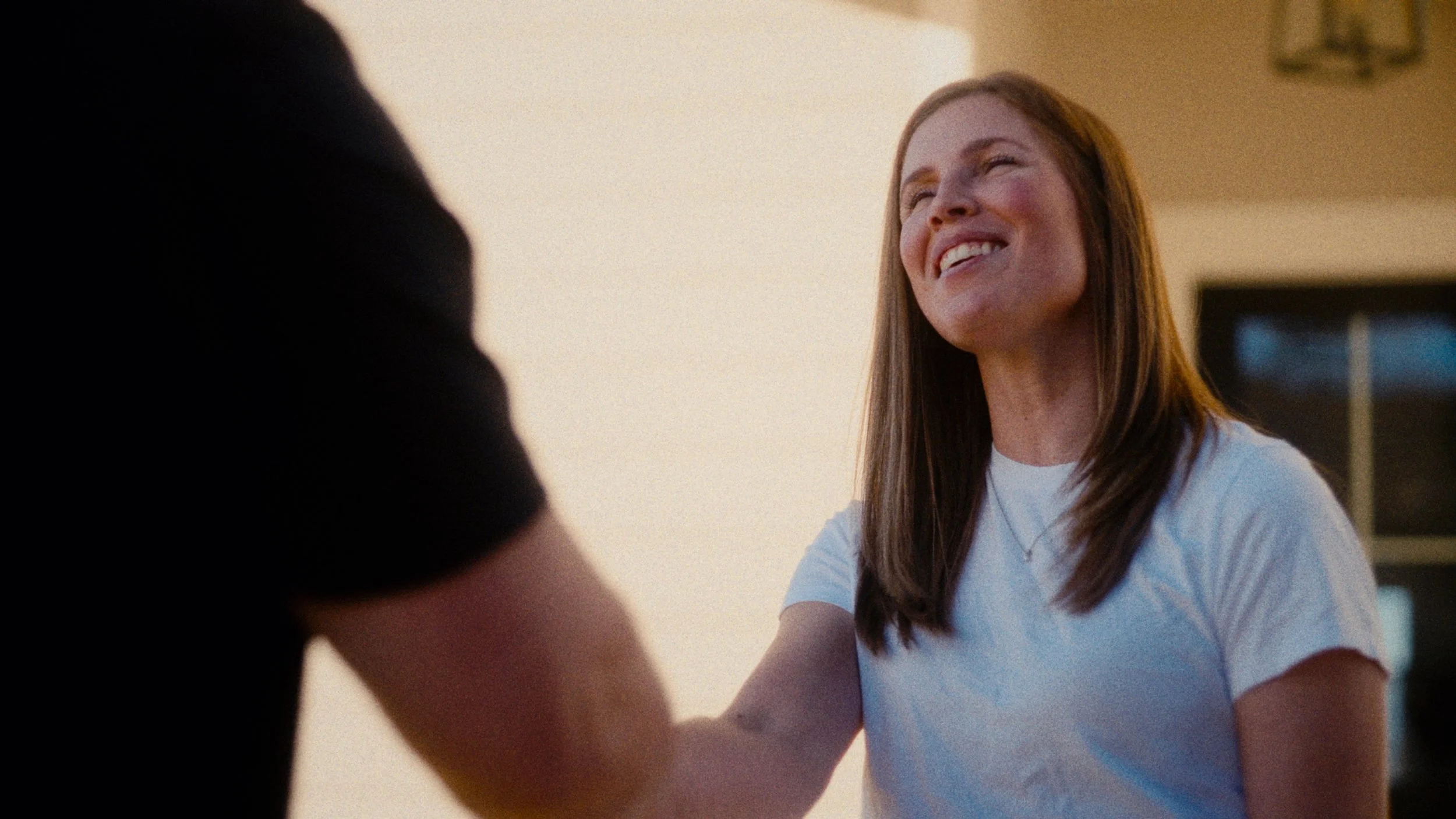 A woman with long brown hair smiling and shaking hands with another person. She is wearing a white t-shirt and a necklace. The background appears to be a home or office setting.