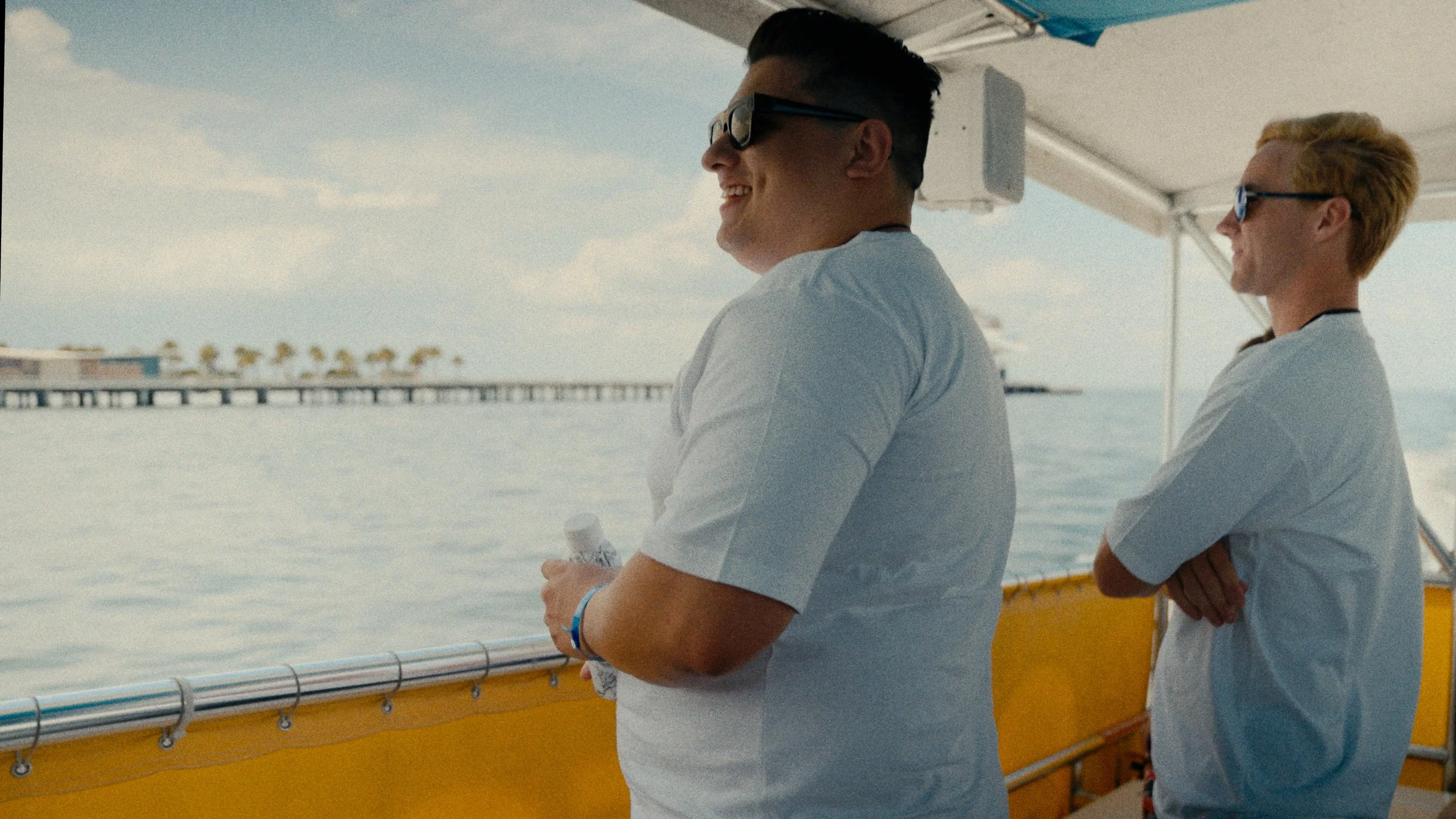 Two smiling men with sunglasses on a boat, standing by the railing, with water and a pier in the background.