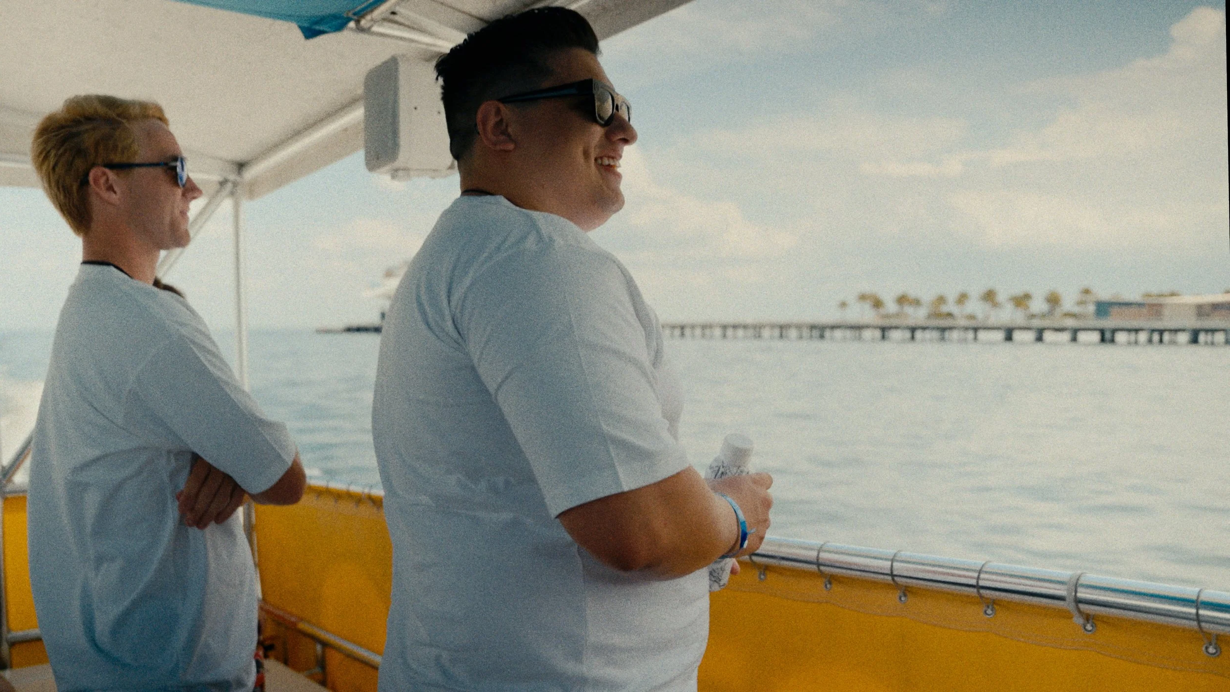 Two people standing on a boat overlooking the water, with a pier and shoreline in the background, smiling and looking at the scenery.