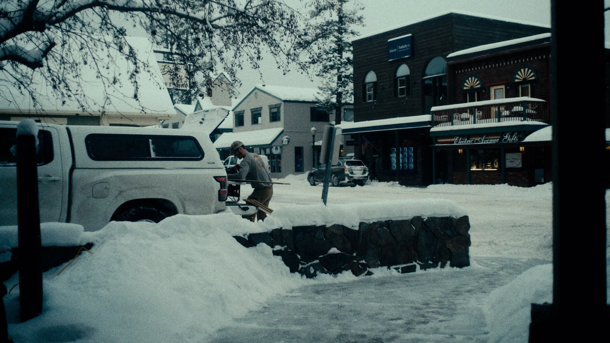 Snow-covered street with buildings and parked cars, one person shoveling snow near a truck, overcast sky, and snow on rooftops.