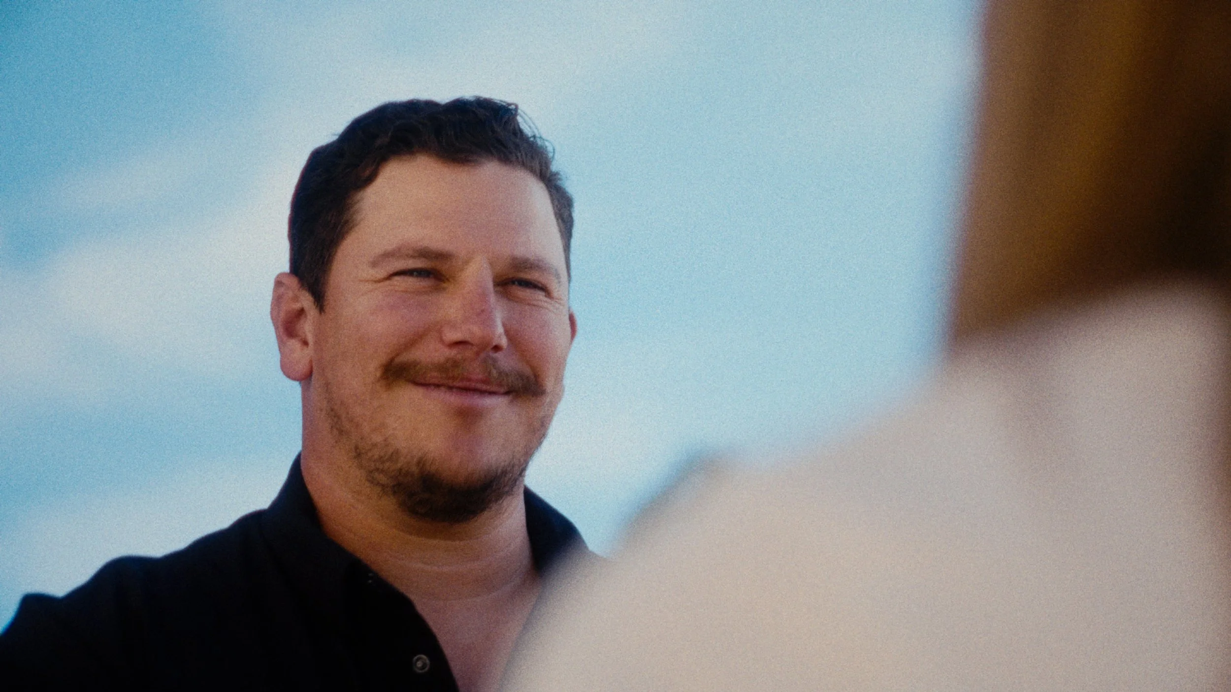 A man smiling outdoors with a blue sky background, sunglasses on his forehead, wearing a black shirt.
