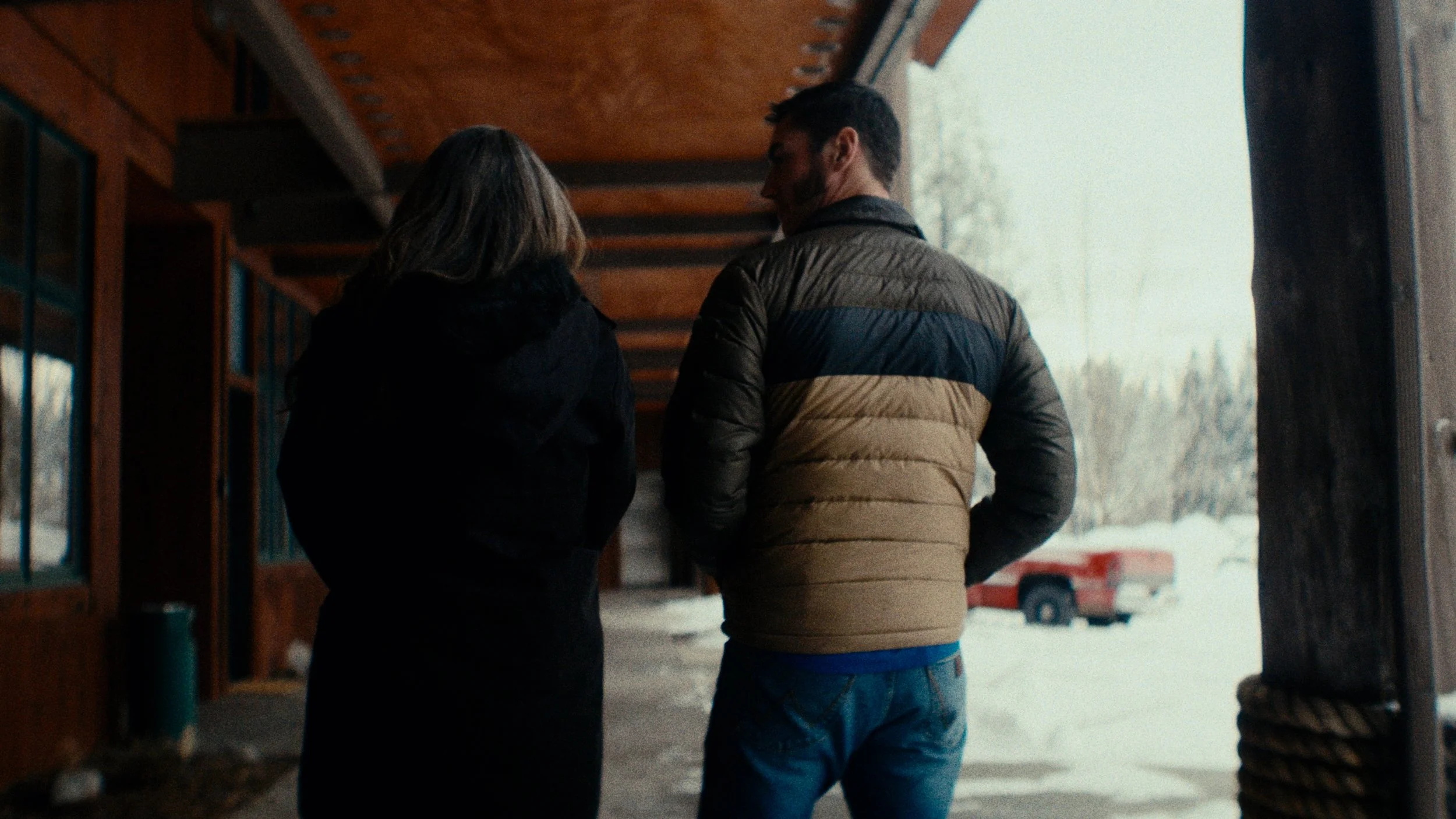 Two people walking away from the camera inside a wooden structure during winter, with snow outside and a red truck visible in the background.