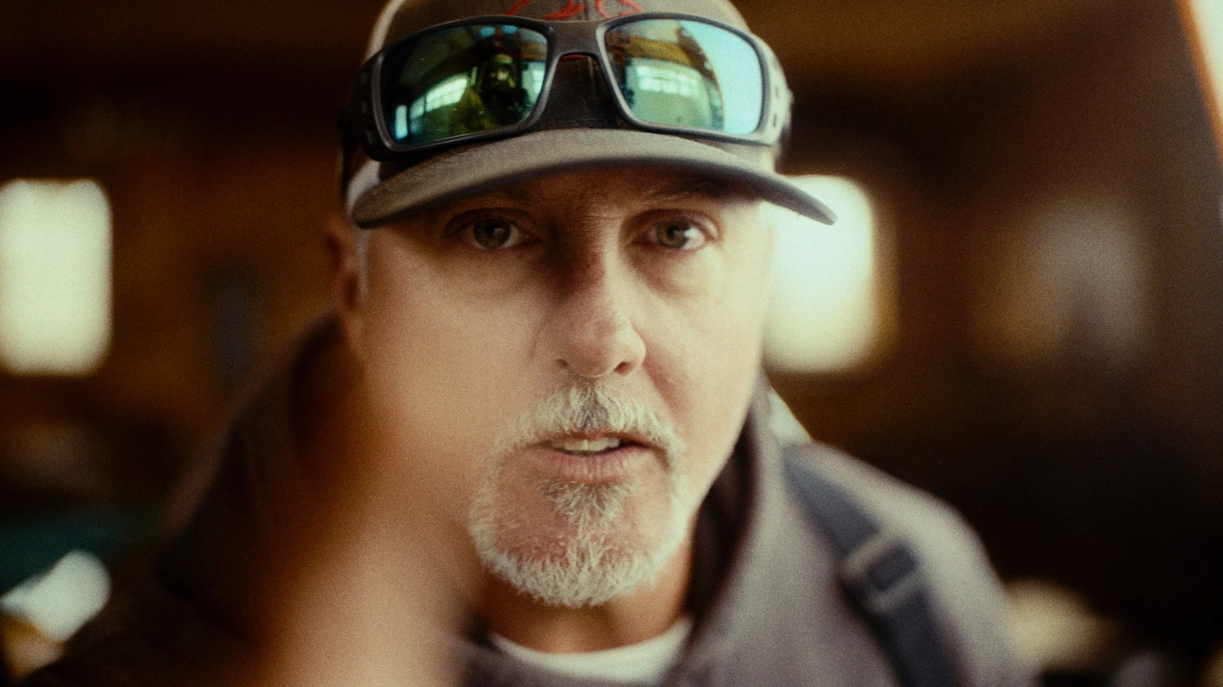 Close-up of an older man with a white beard wearing a camouflage cap and sunglasses, looking directly at the camera.
