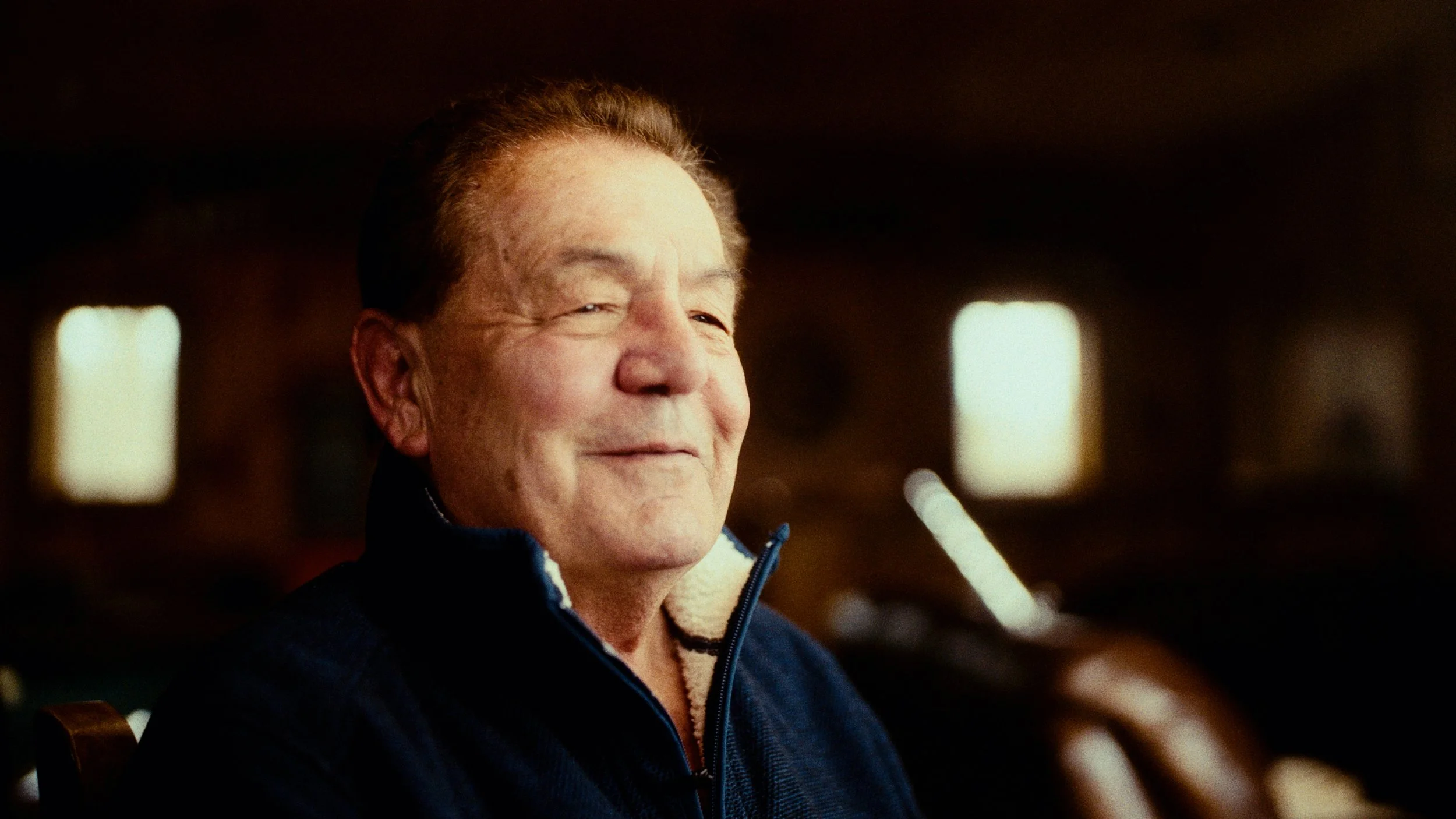 A man with short light brown hair smiling, wearing a blue jacket with a zipper, sitting indoors with blurred windows in the background.
