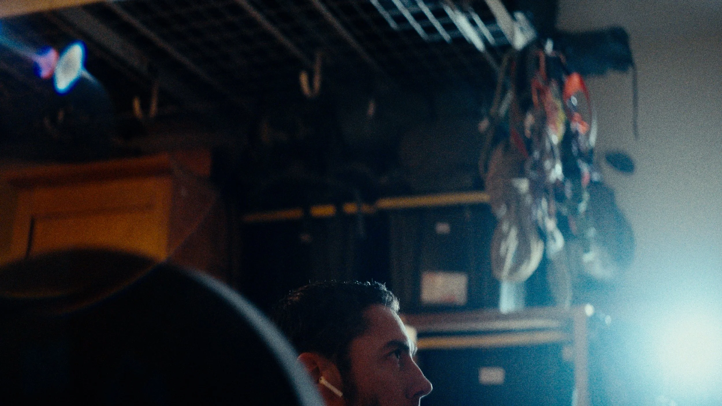 Side profile of a man in a dark room with hanging climbing shoes and gear above him, illuminated by bright light from the right.