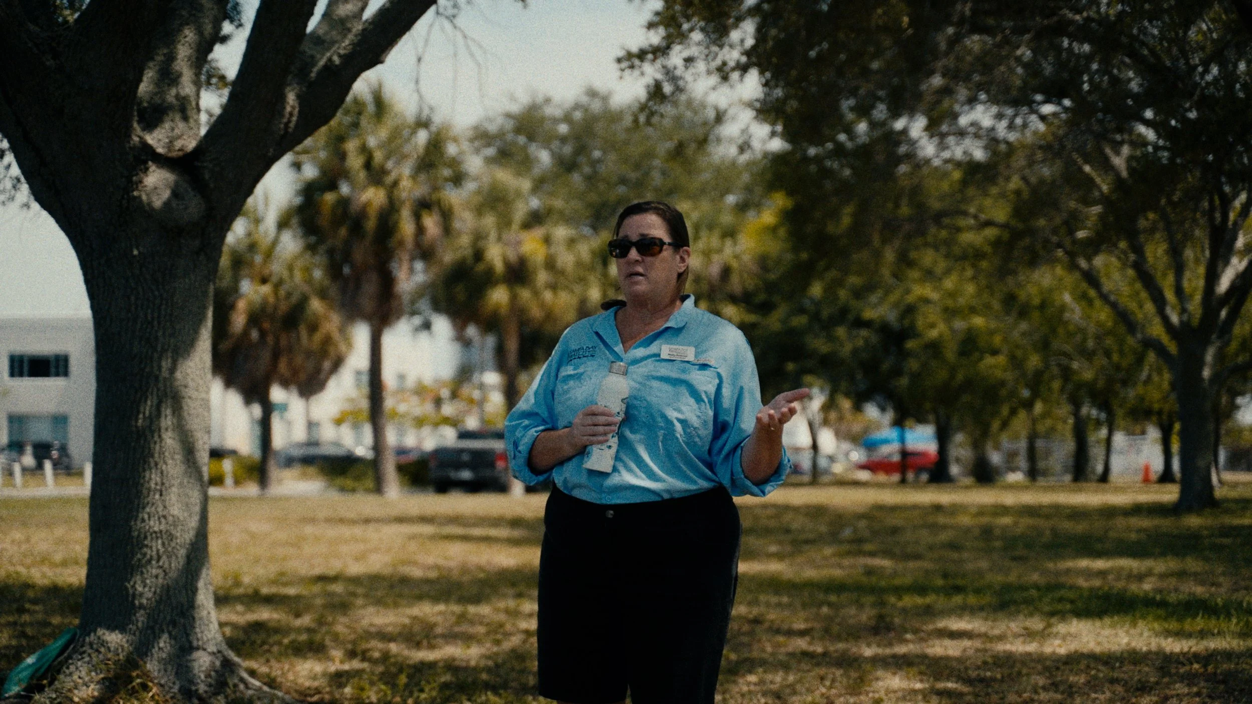 A woman in a light blue uniform shirt and black skirt standing outdoors under a tree, holding a water bottle, with a background of trees, cars, and buildings during daytime.