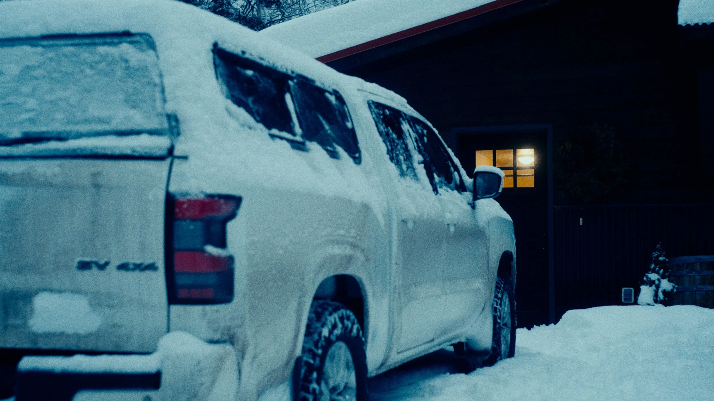 A white pickup truck parked in driveway covered with snow, in front of a house with a lit window.