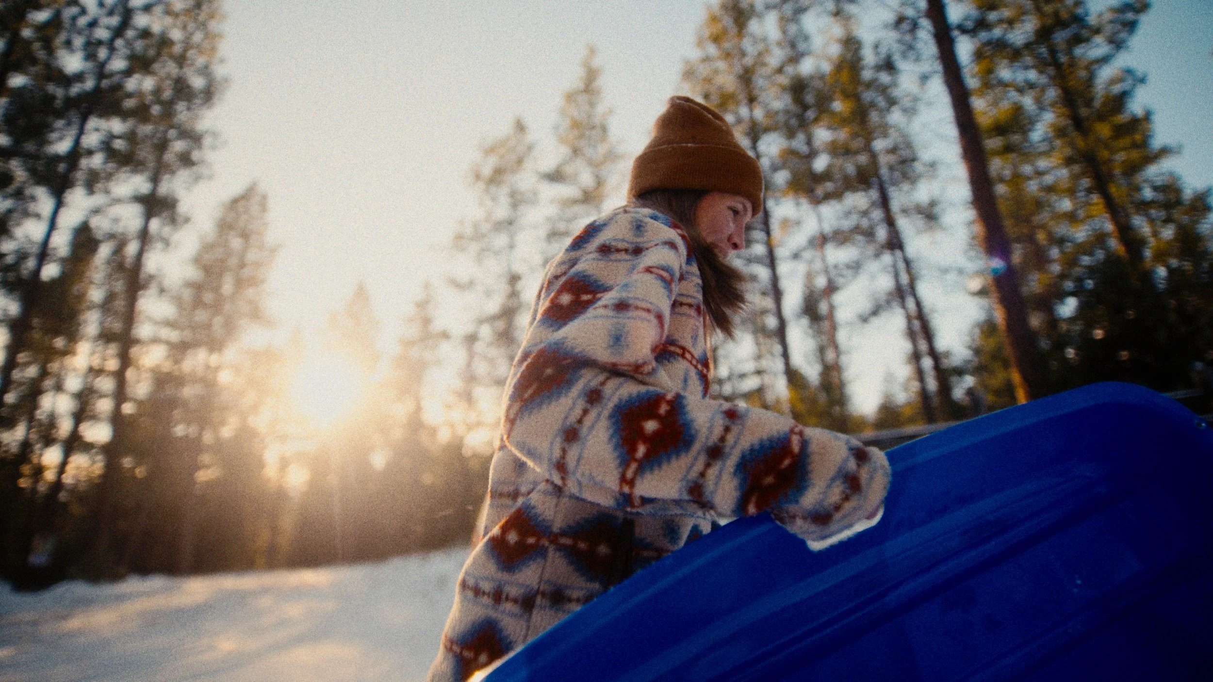 A woman wearing a patterned jacket and a brown beanie is outdoors in a snowy forest, holding a blue object, with sunlight filtering through trees in the background.