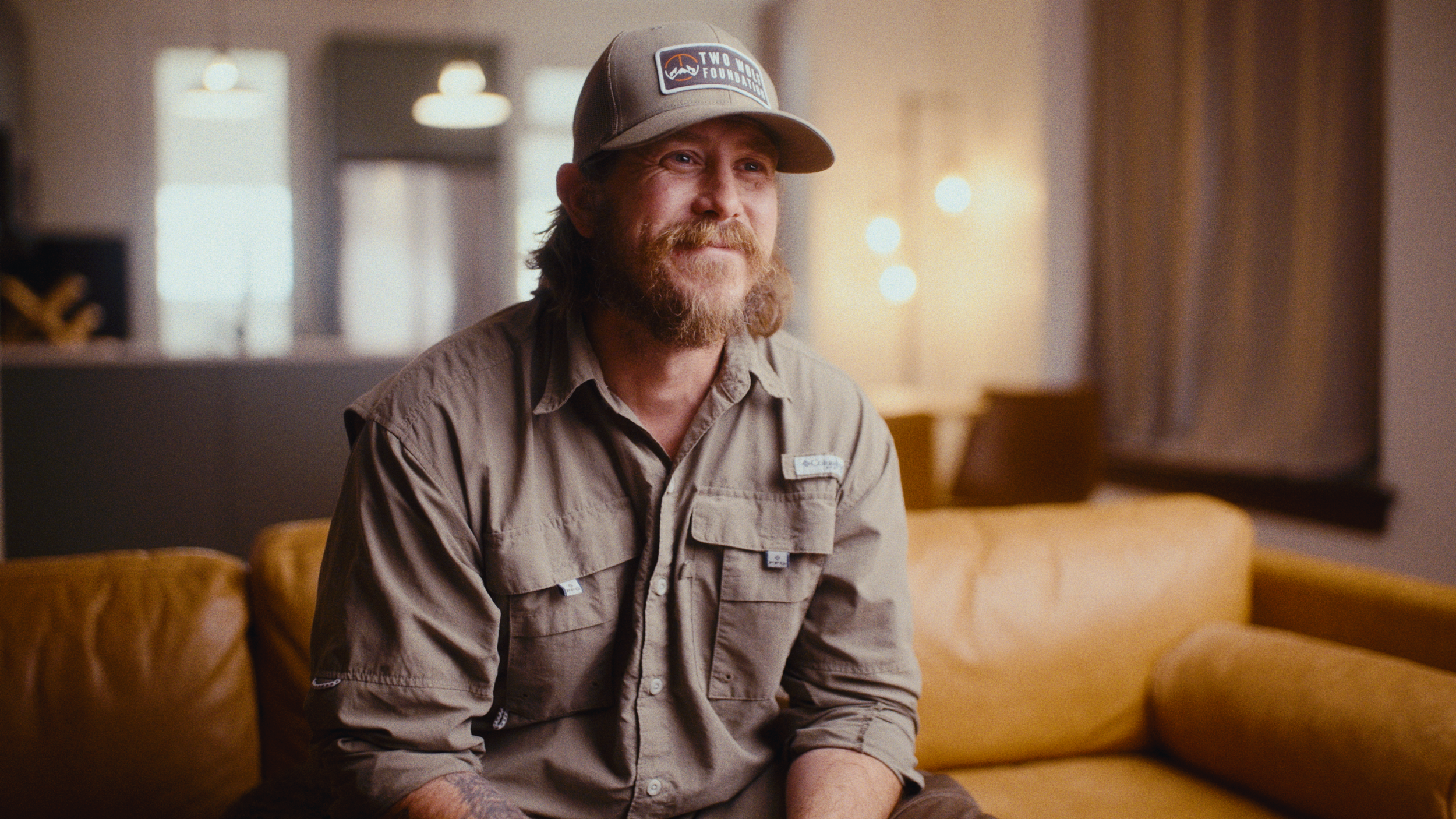 A man with a beard and long hair wearing a beige shirt and a cap that says 'Two Wood Foundation' is sitting on a yellow couch in a warmly lit room.
