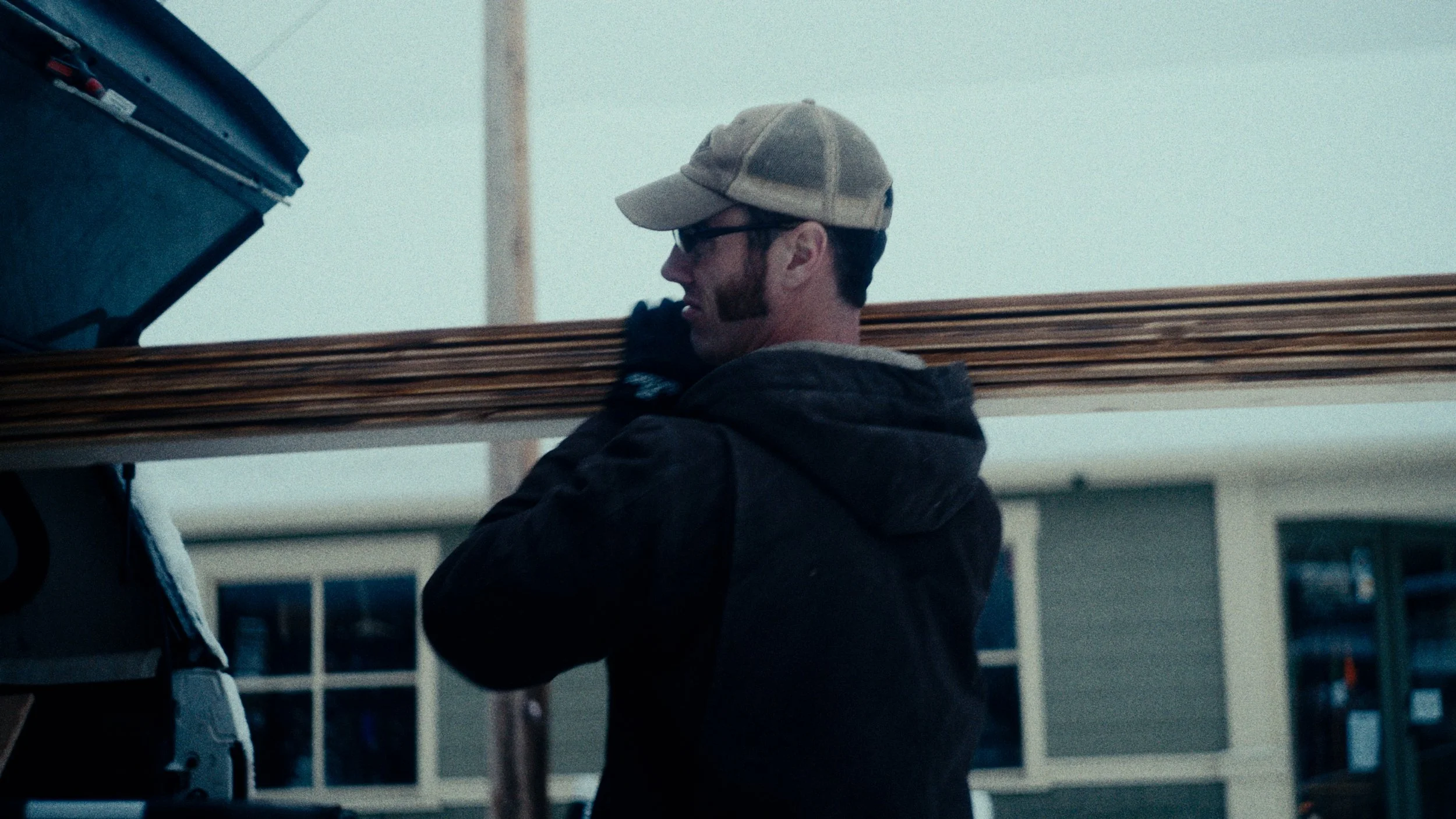 A man wearing a baseball cap, sunglasses, and a dark hoodie is carrying wooden planks on his shoulder outside in front of a house.