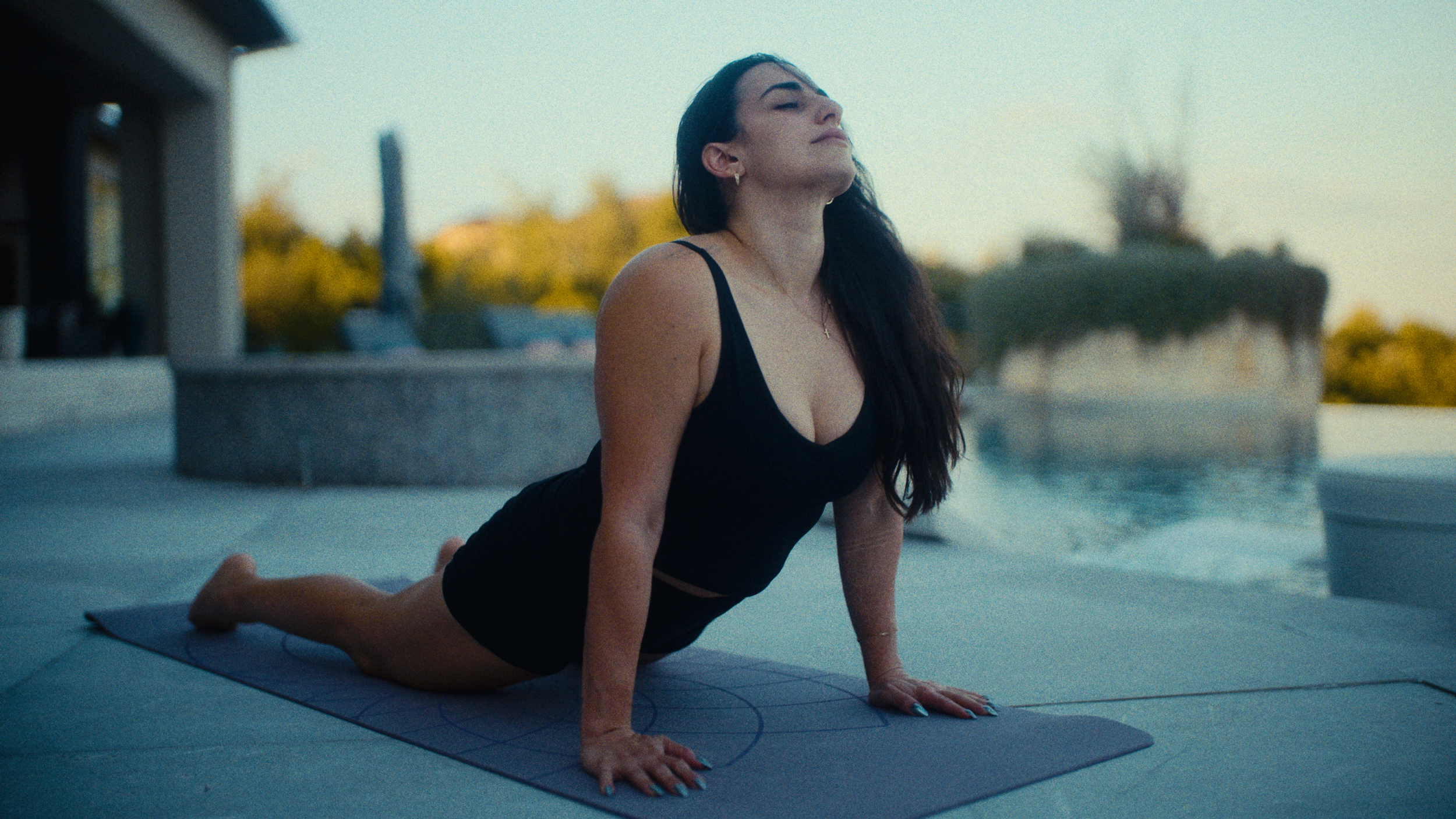 Woman practicing yoga outdoors near water, in a plank position on a yoga mat with her eyes closed and a serene expression.