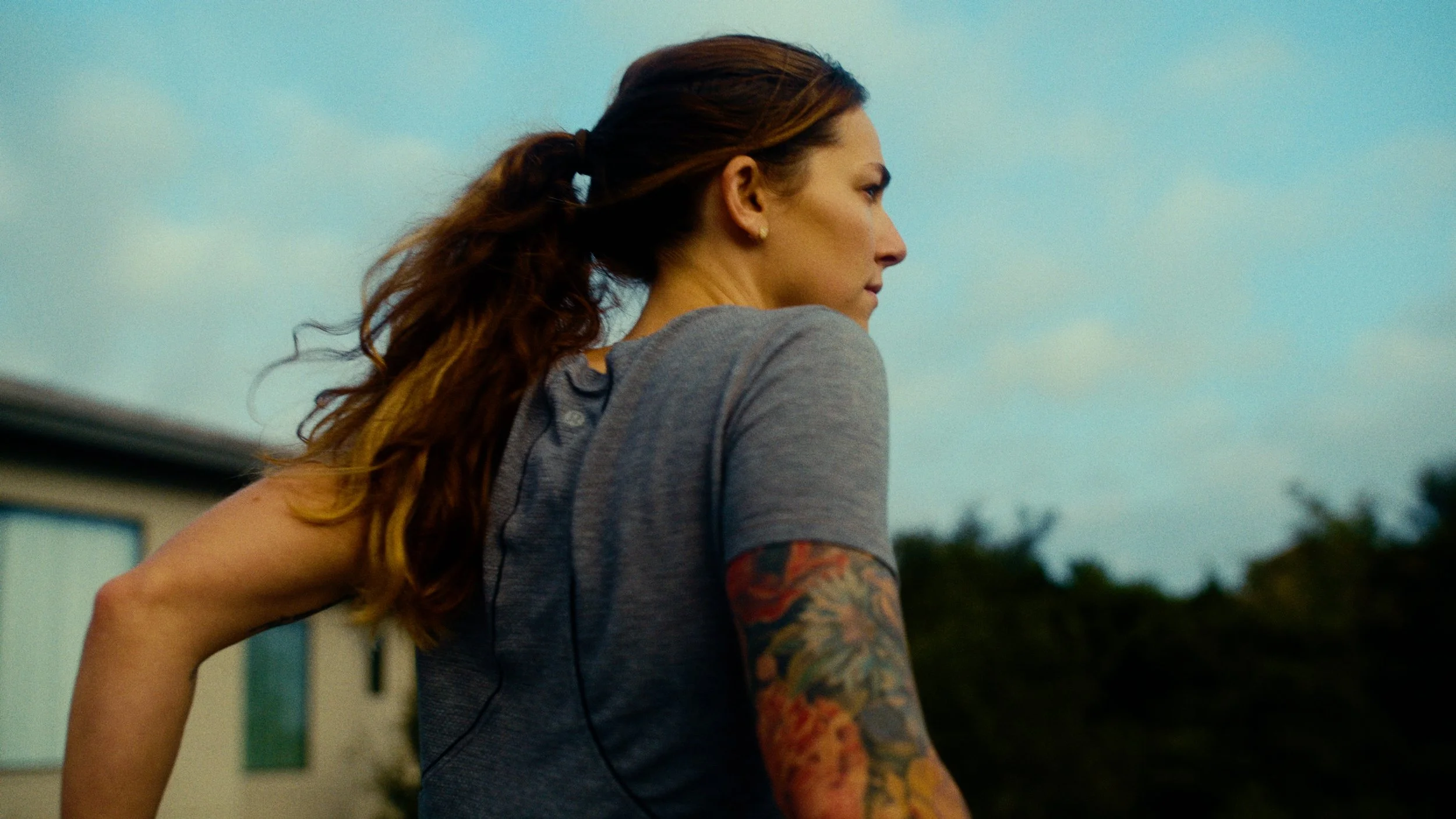A woman with long, wavy hair in a ponytail, wearing a gray t-shirt and earphones, stands outdoors against a cloudy sky and trees in the background.