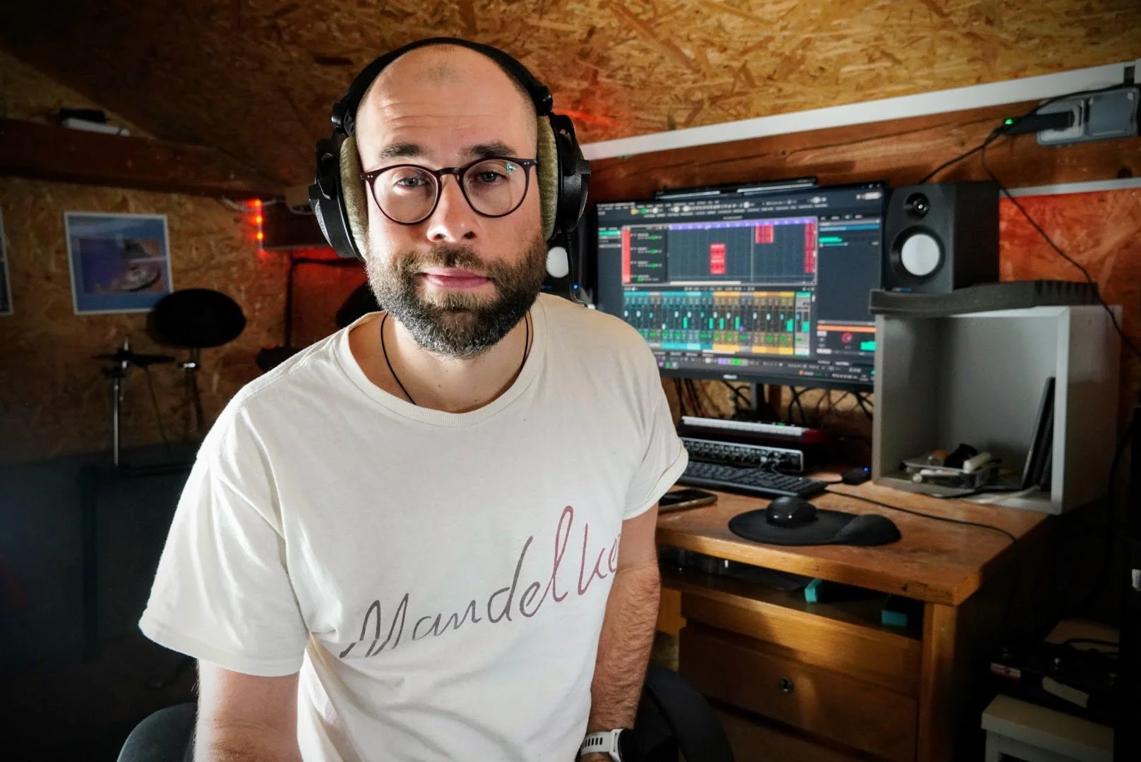 Ein Mann sitzt in einem Tonstudio vor einem Computer mit Musiksoftware, trägt Kopfhörer und Brille, im Hintergrund sind Musikinstrumente und Wanddekoration sichtbar.
