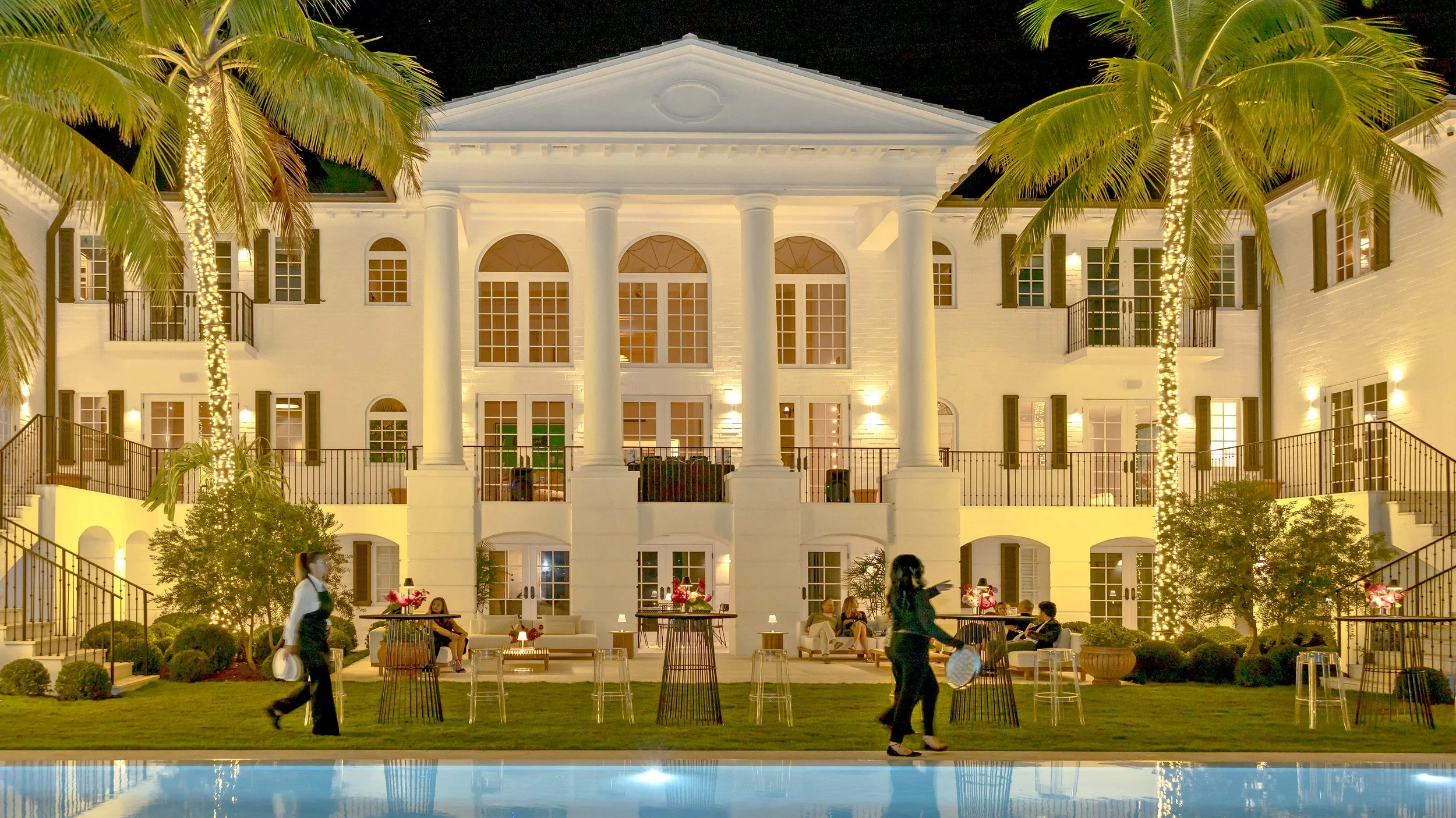Night view of an Artsy event at an elegant white mansion in Miami during Art Basel, featuring tall columns, lit windows, a reflection fountain, and outdoor dinner seating.