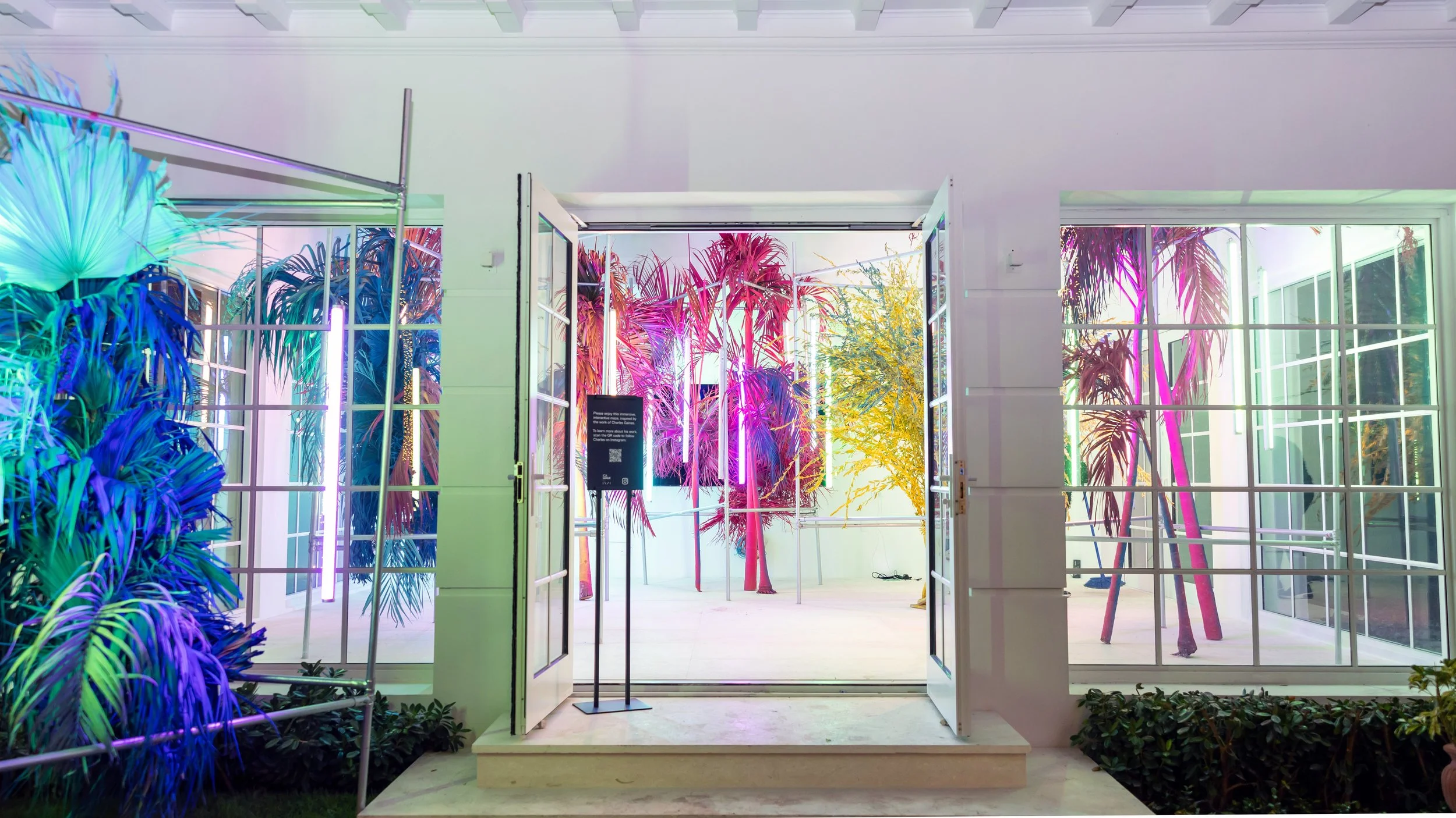 An immersive artsy event installation featuring neon-lit tropical foliage, palm trees, and modern lighting seen through open double doors.