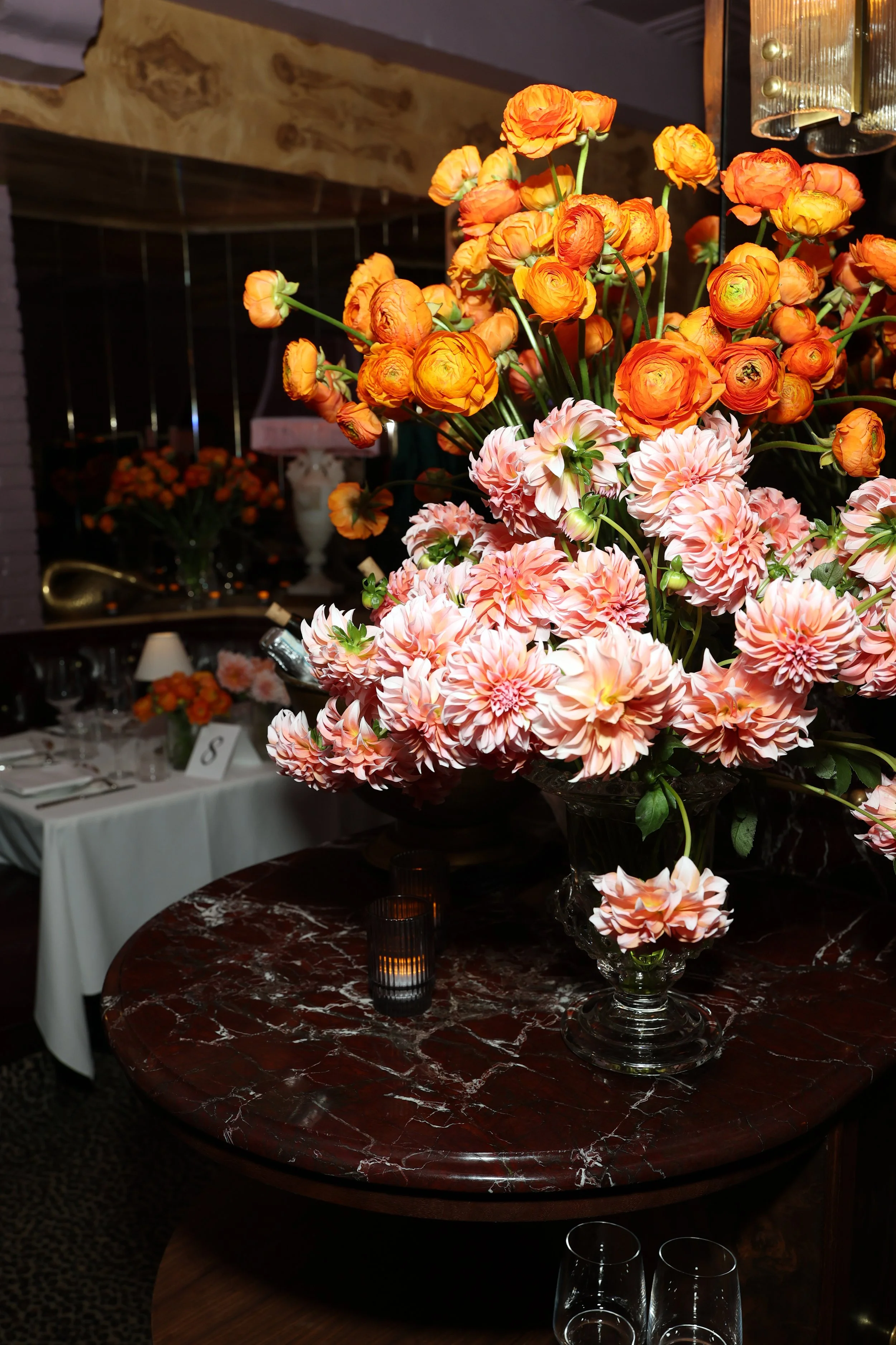A floral centerpiece with vibrant orange ranunculus on a red marble table, illuminated by candles in an elegant event setting.