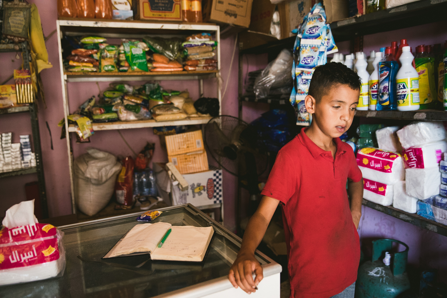  A young boy standing in his family’s small shop inside the Shatila camp. As most of refugees lack a regular source of income many products are purchased on loan, and ledgers keep track of the community debts. 