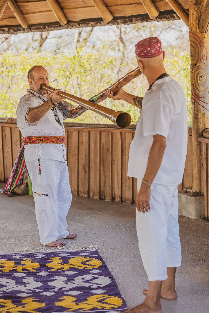 Two people in white traditional attire play didgeridoos indoors near a wooden railing.