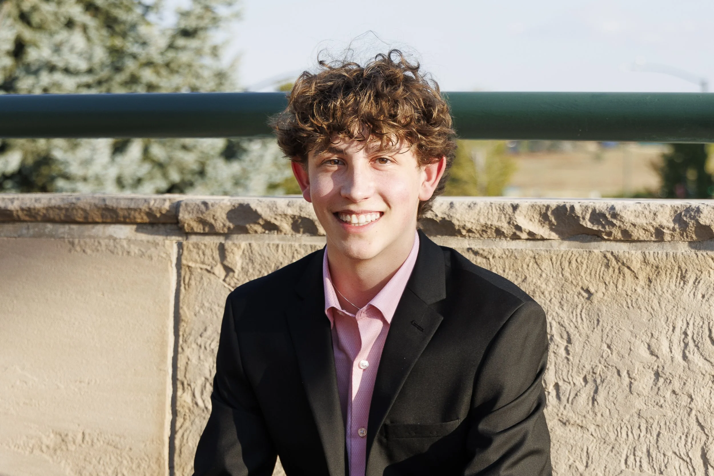 Young person with curly hair smiling outdoors in a black suit and pink shirt