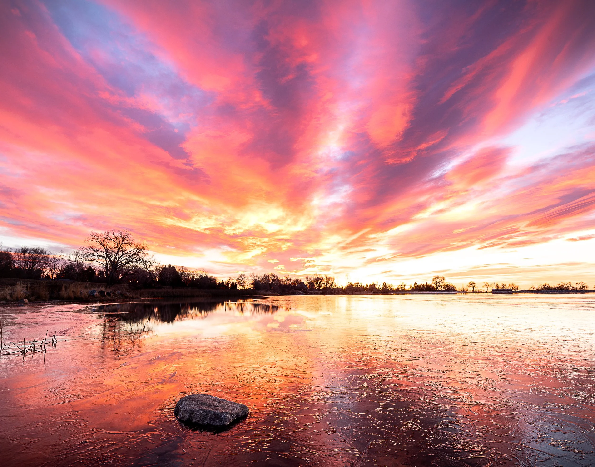A vibrant sunset with pink, orange, and purple clouds reflected on a calm lake with a rocky shore and silhouette of trees.