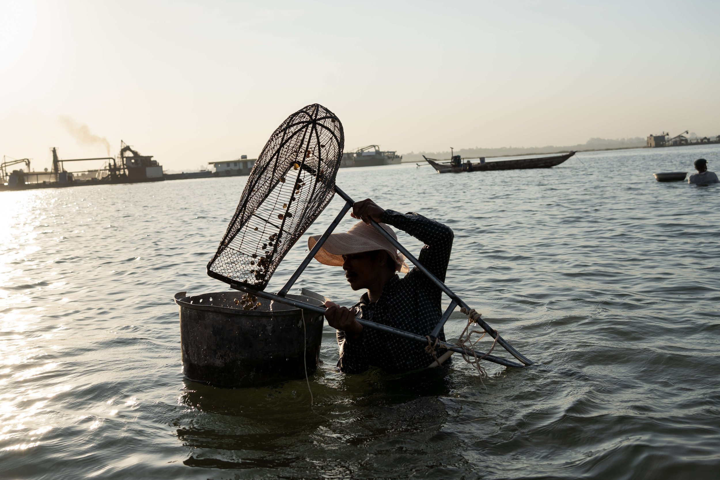  A fisher, who requested anonymity due to fear of retribution from authorities and the well-connected sand mining companies operating in the area, catches snails in the Mekong as sand mining barges work in the background. Before the barges arrived, h