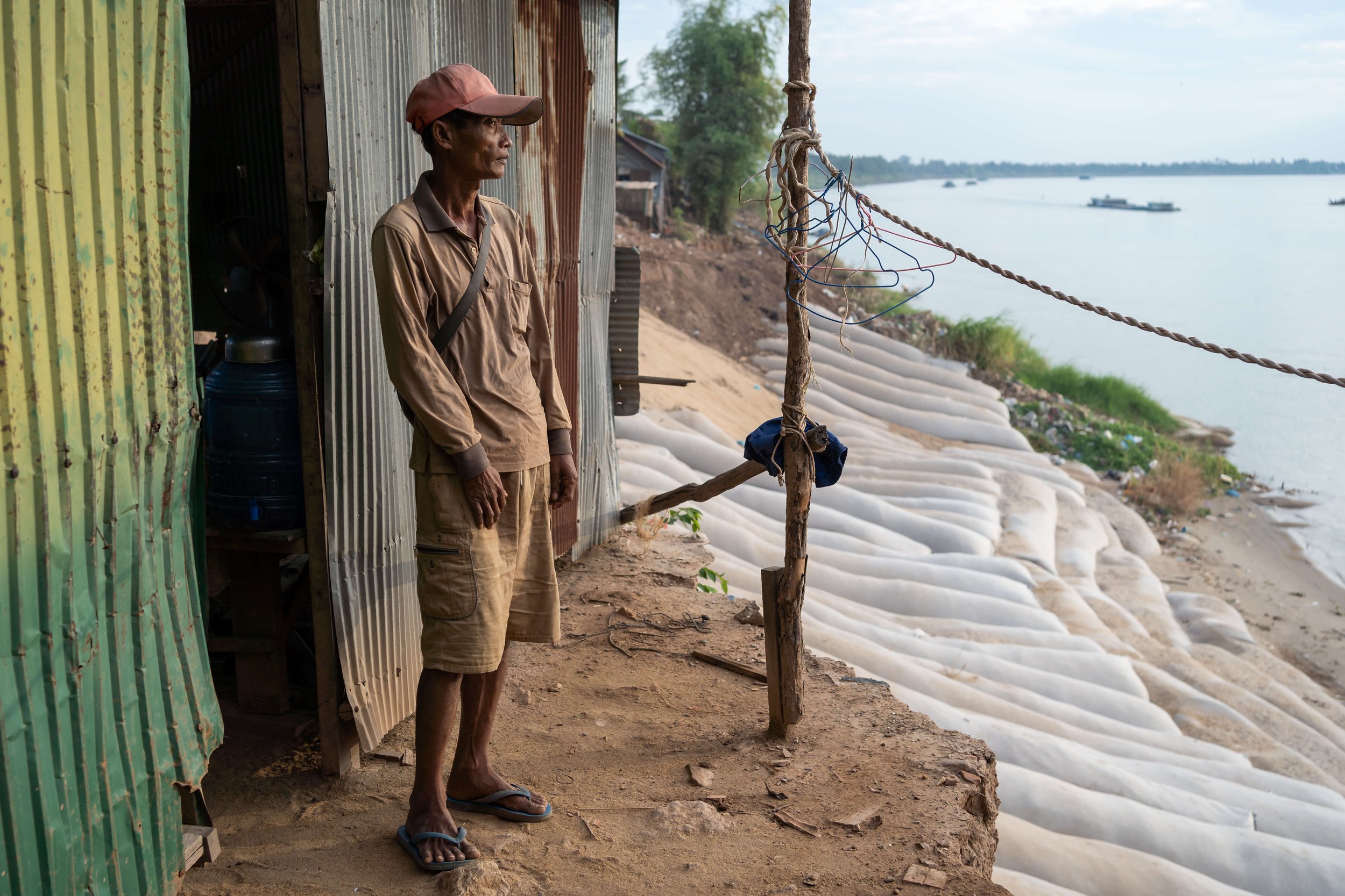 In Chhai Heng, a remorque driver, stands for a portrait at his house next to one of two river bank collapses that happened in 2021 along the same road in Rokar Koang. In the distance, sand barges deliver sand to the capital Phnom Penh. Like many oth
