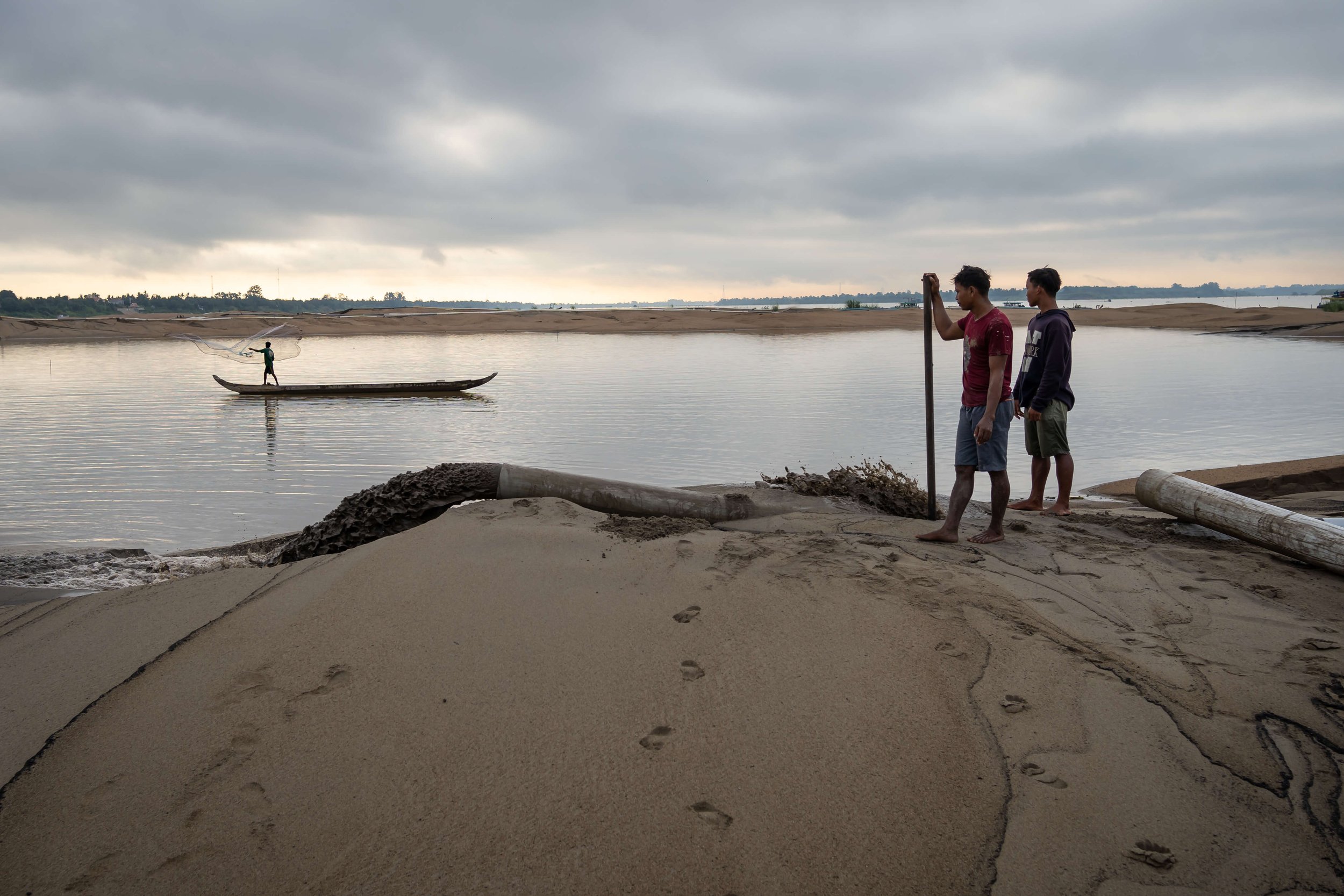  Sand mining workers look on as a fisher casts his net into a temporary lake created by one of Phnom Penh’s latest major land reclamations along the Mekong river. As the city continues to grow, so too does the thirst for sand, leading to an uncertain