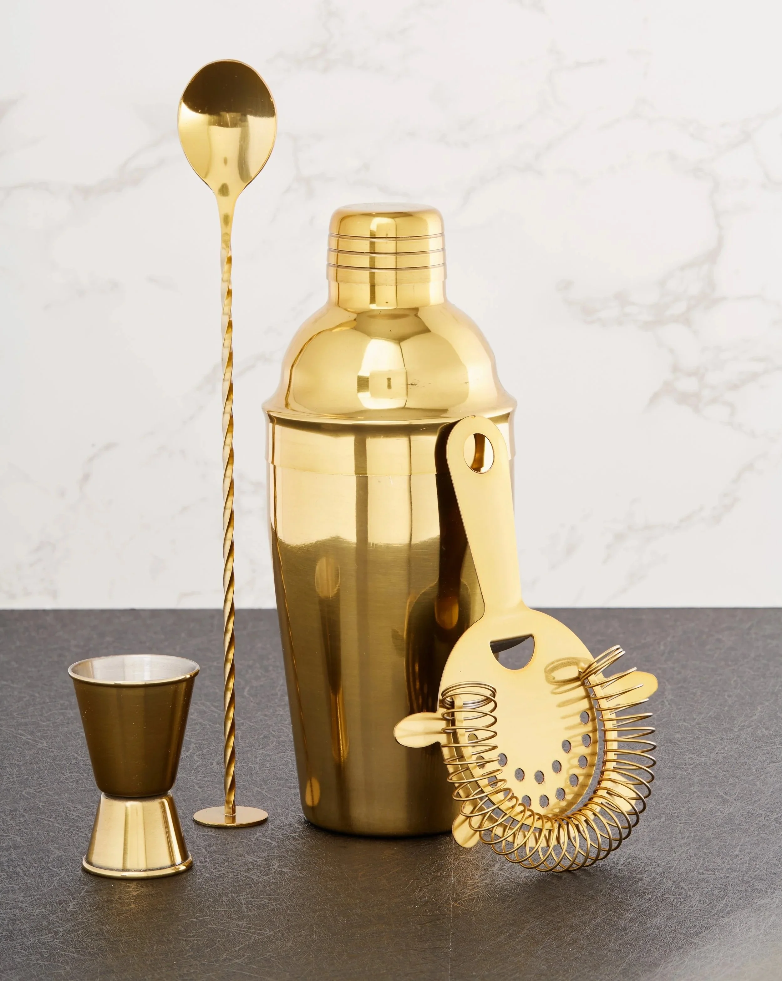 Gold-tone cocktail shaker and bar tools arranged against a marble backdrop.