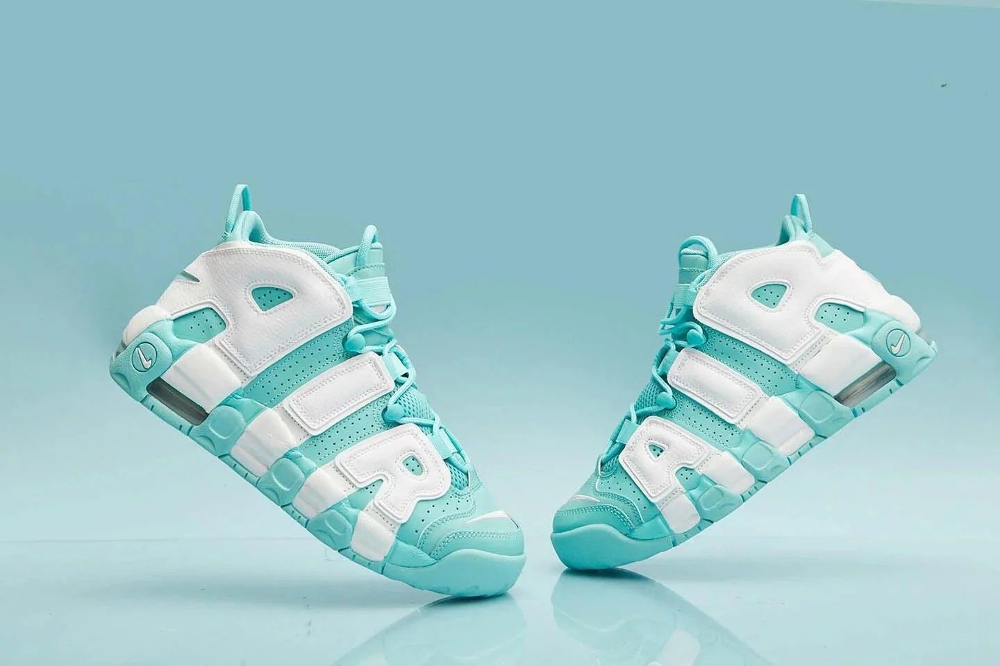 Pair of white and turquoise Nike Air More Uptempo sneakers photographed against a matching turquoise background.