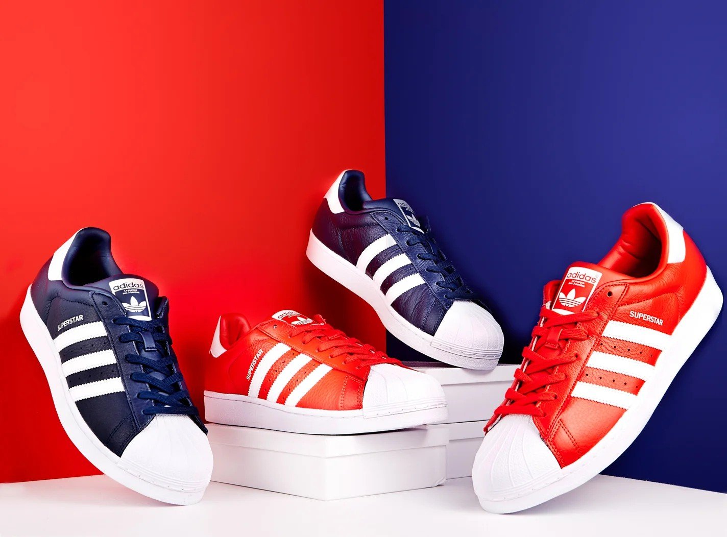 Four Adidas Superstar sneakers, in navy and red colorways, displayed on white platforms against a red and blue background — commercial product photography.