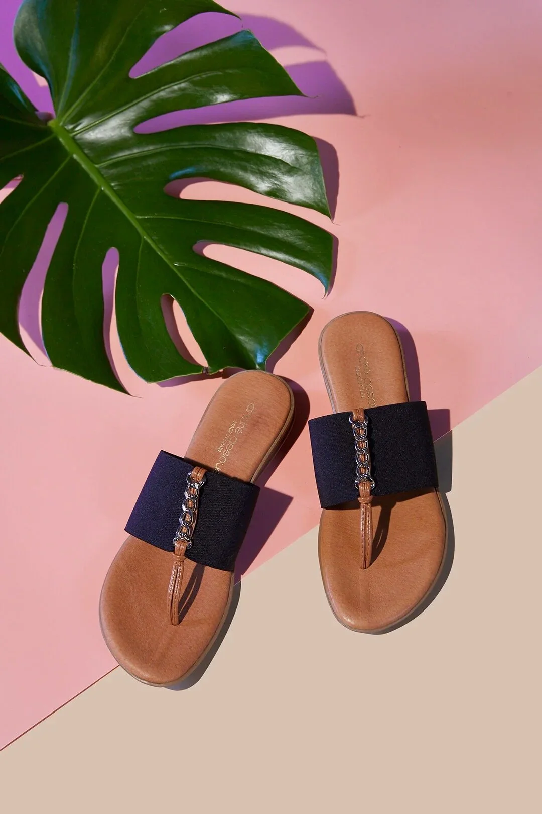 Pair of tan slide sandals with black straps and chain detail on a pink and beige background next to a green monstera leaf.