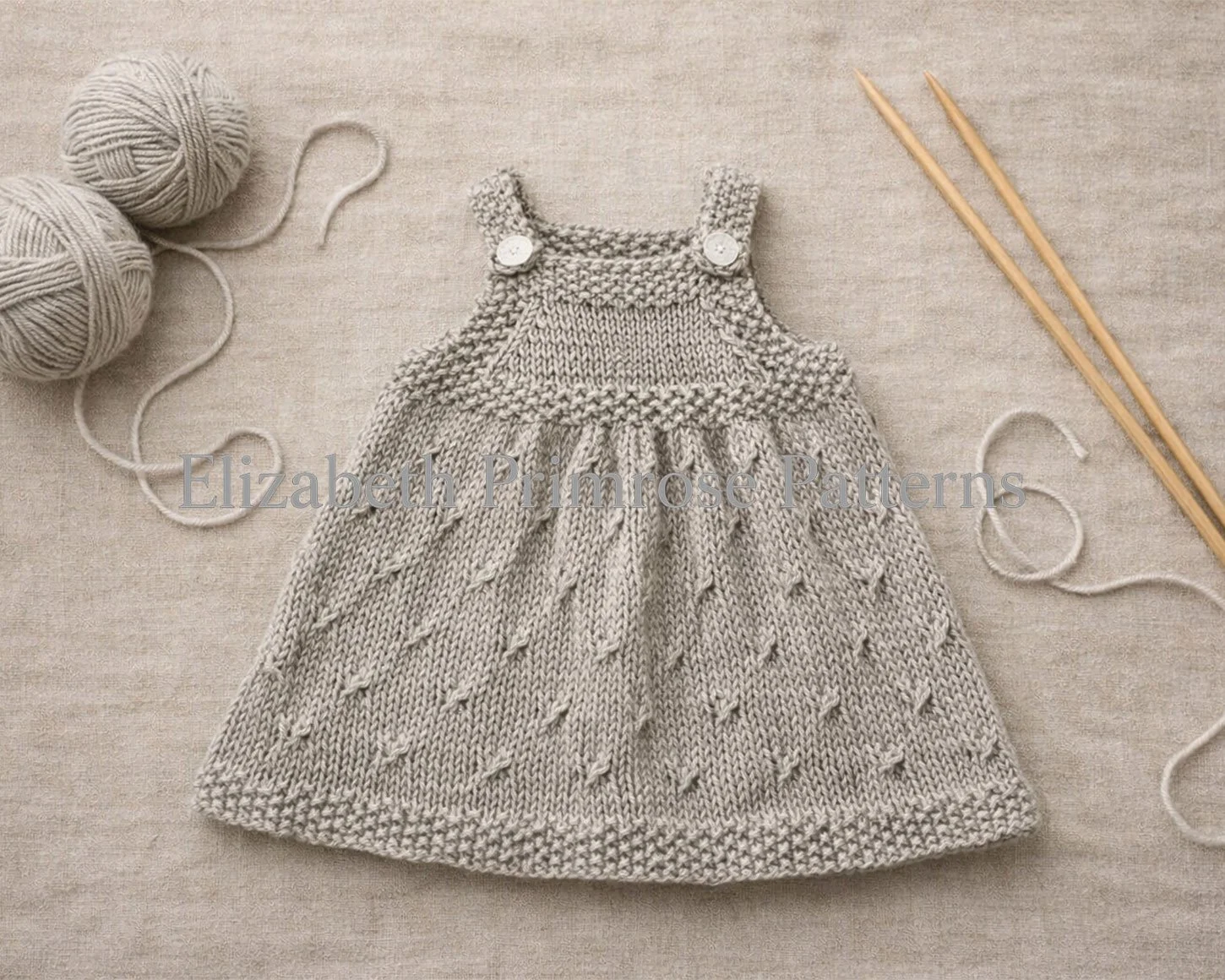 Beige hand knitted dolls pinafore laid flat on beige fabric, with textured skirt, buttoned straps, matching yarn balls and wooden needles beside it.