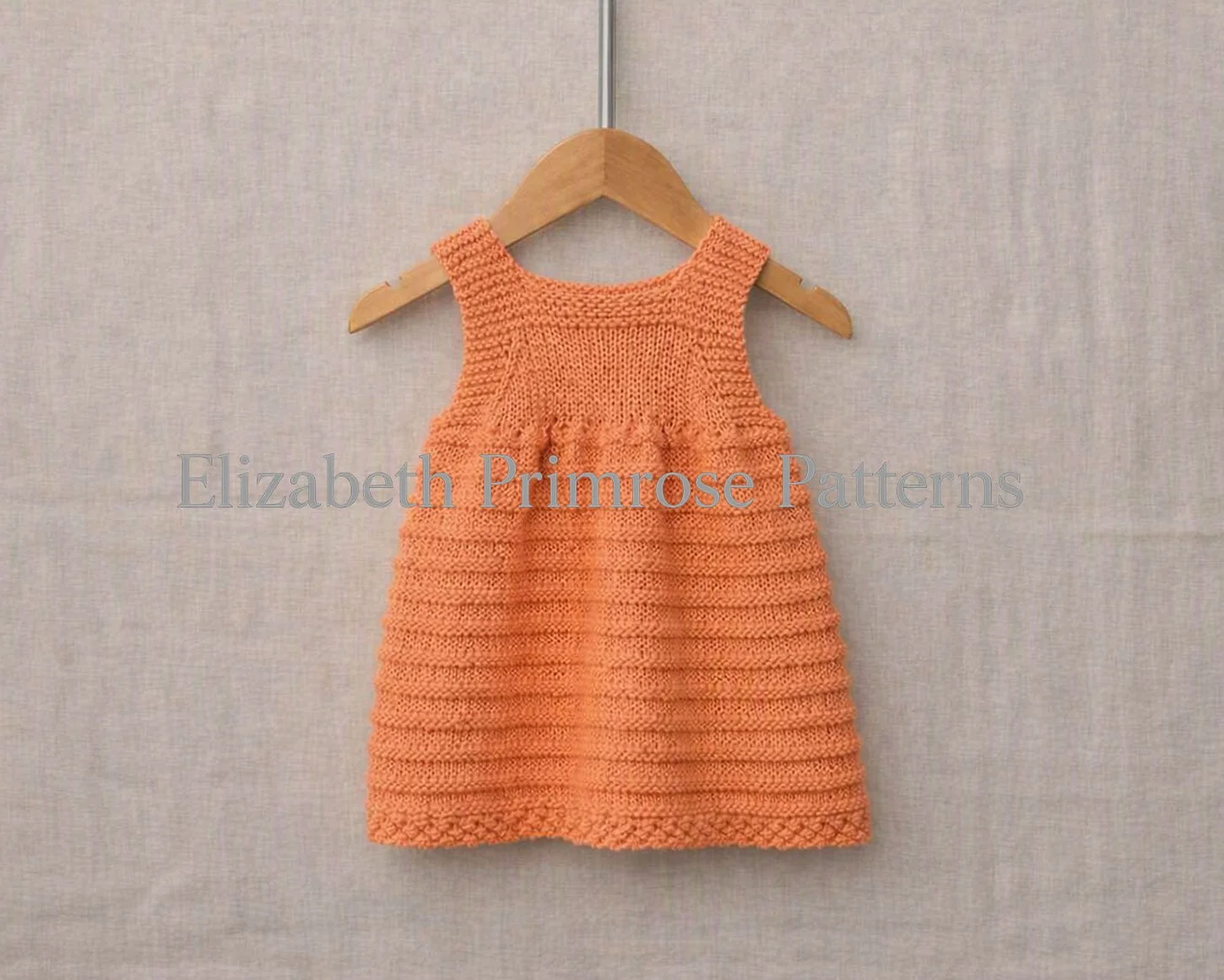 Back view of hand‑knitted peach baby pinafore with textured ridges and bobbles displayed on a hanger over a neutral fabric backdrop.