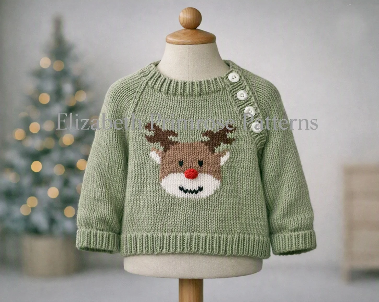 Sage green hand knitted raglan jumper with reindeer motif and buttoned shoulder, displayed on wooden mannequin in softly lit Scandinavian children’s room.