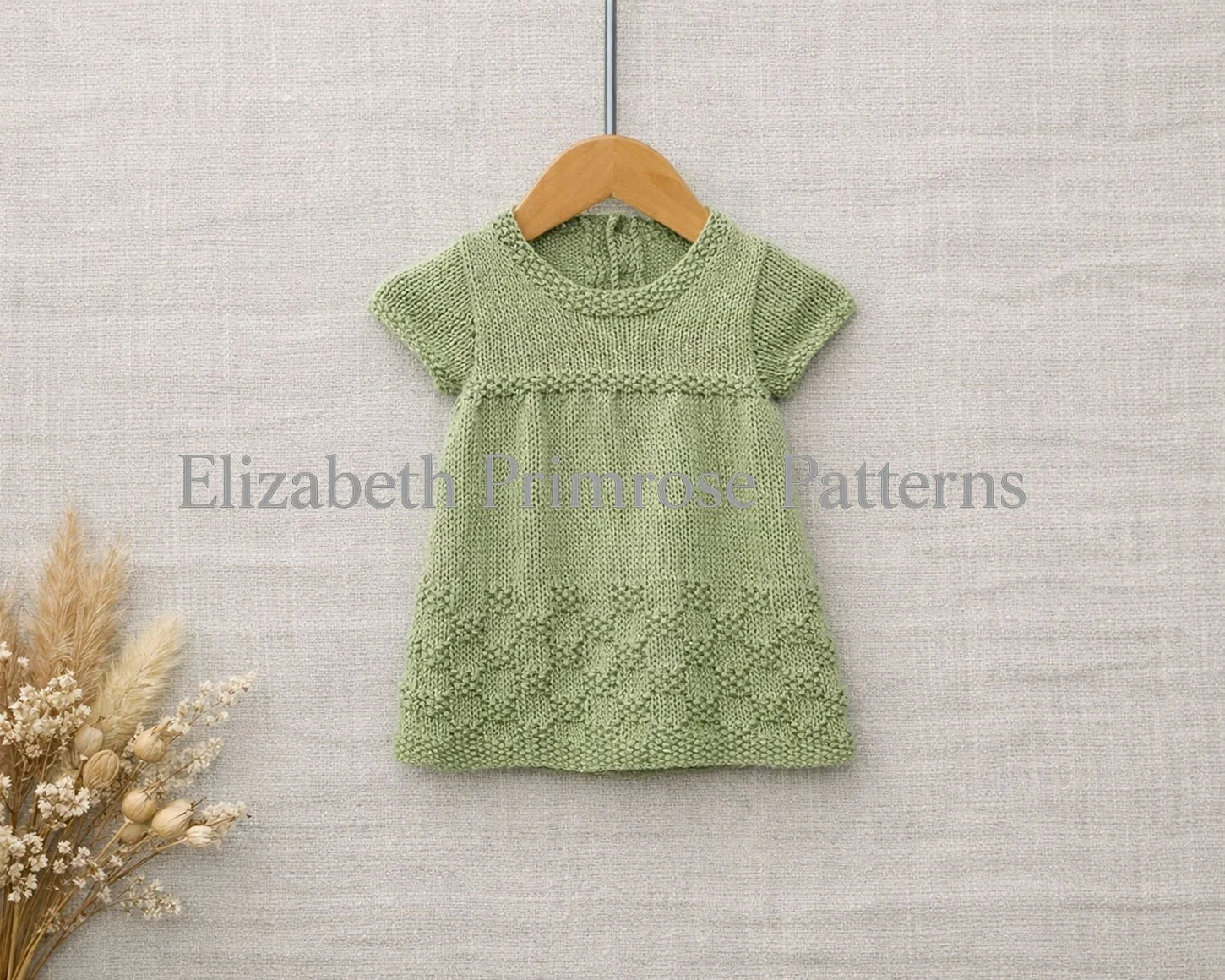 A hand‑knitted, short-sleeved green children’s dress on a wooden hanger, shown against textured beige linen with dried flowers in the corner.