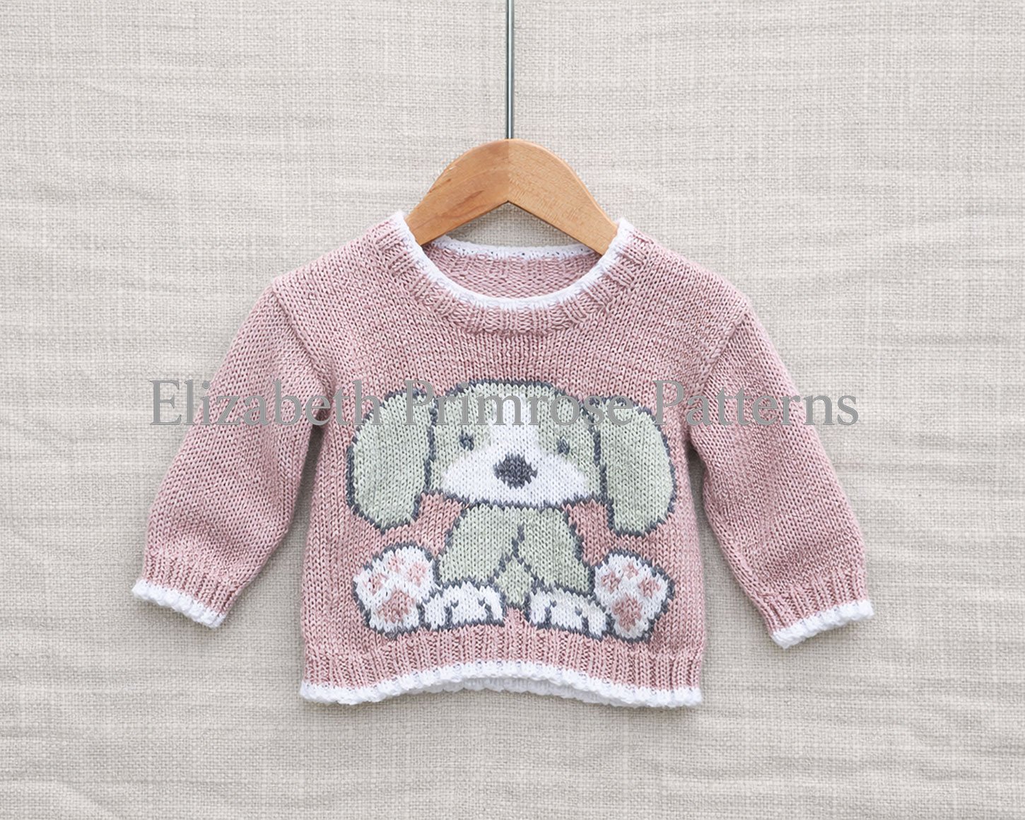 Front view of pink knitted baby jumper with white trim and dog motif, hanging on wooden hanger against linen background.