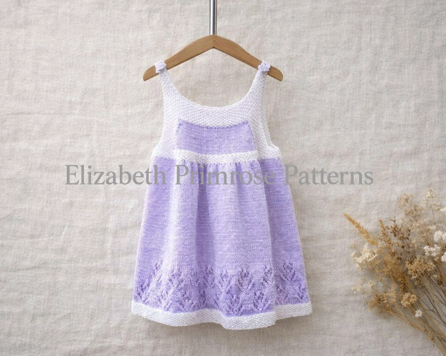 Lavender and white knitted pinafore dress hanging on a wooden hanger against a beige linen backdrop with dried flowers beside it.