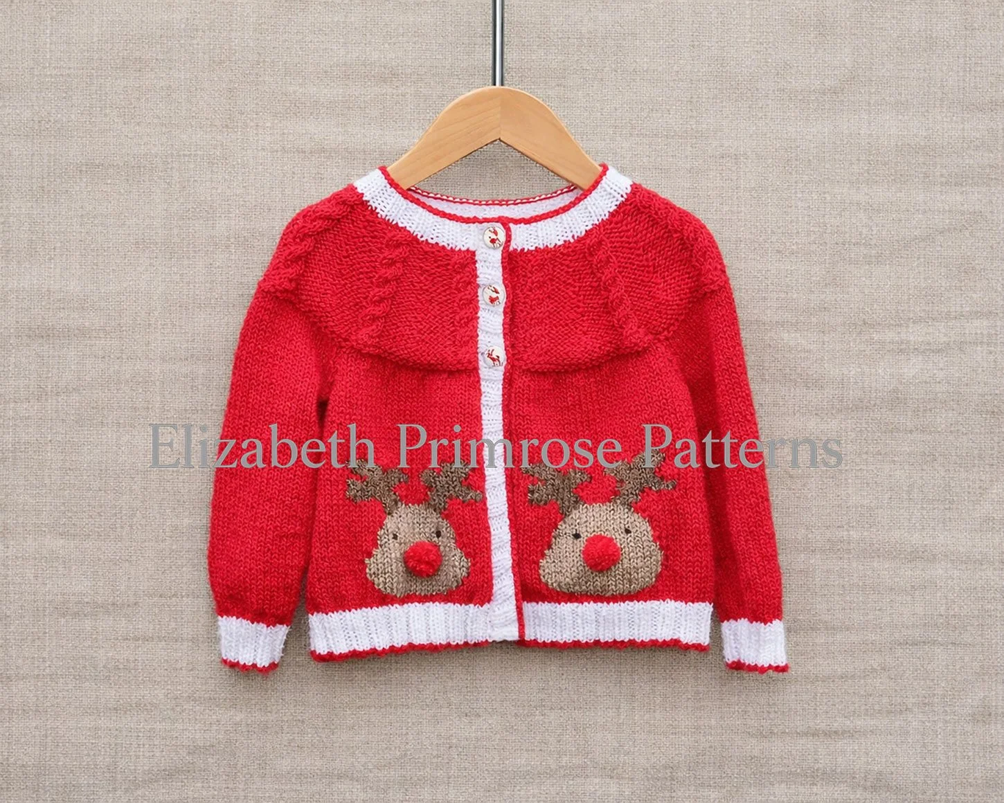 Red children’s Christmas cardigan with reindeer motifs on a wooden hanger against a beige fabric background, showing clear stitches and festive detailing.