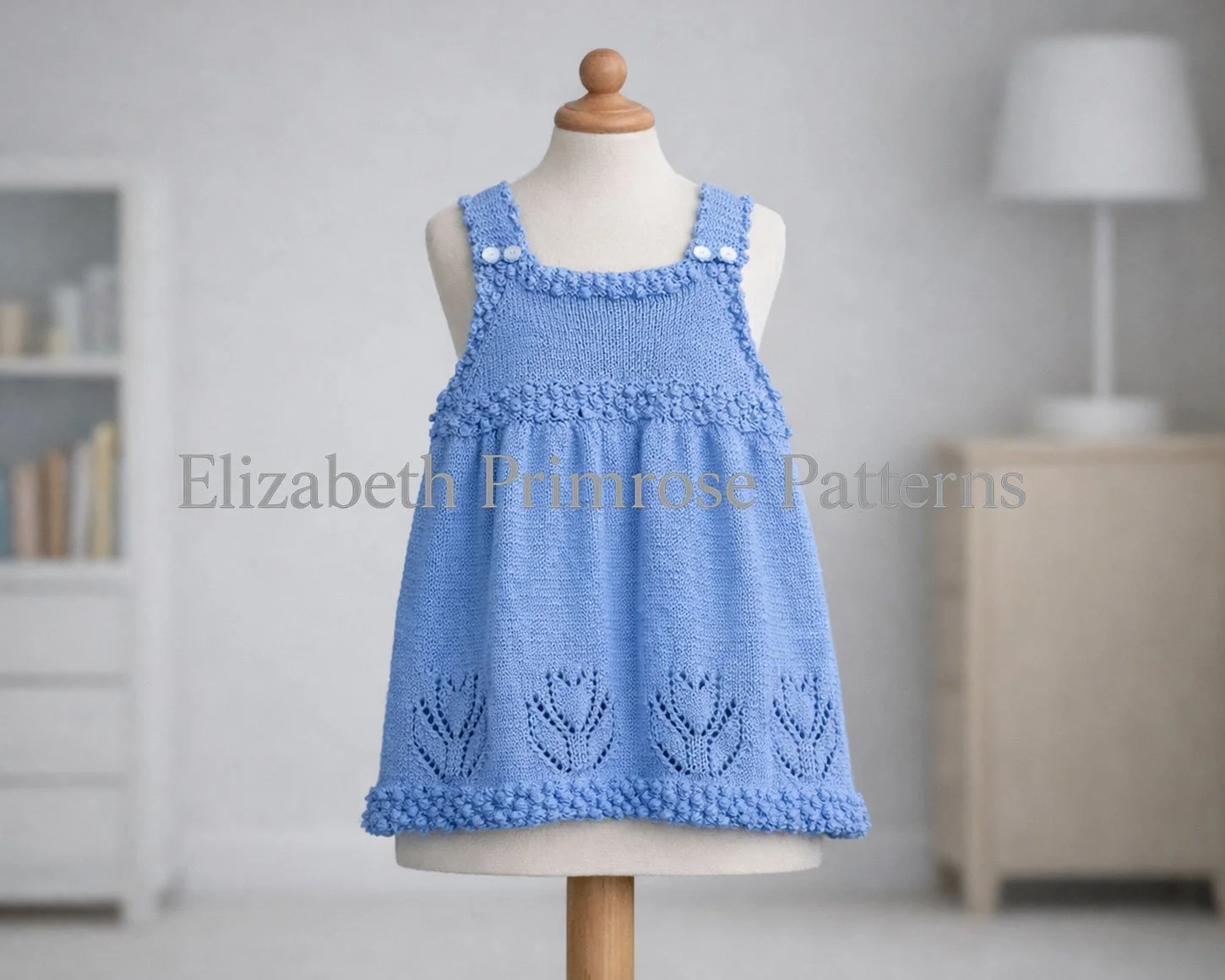 Knitted blue dress with bobble hem and tulip pattern displayed on child mannequin in blurred nursery background with light furniture.