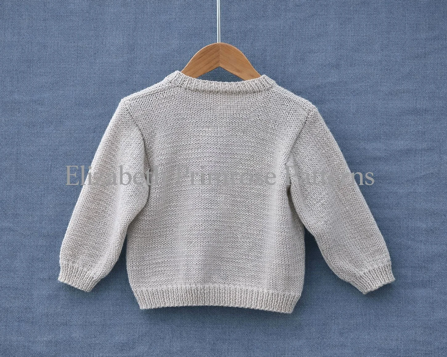 Back view of knitted cream children’s cardigan, hanging on a wooden hanger before a textured blue backdrop.