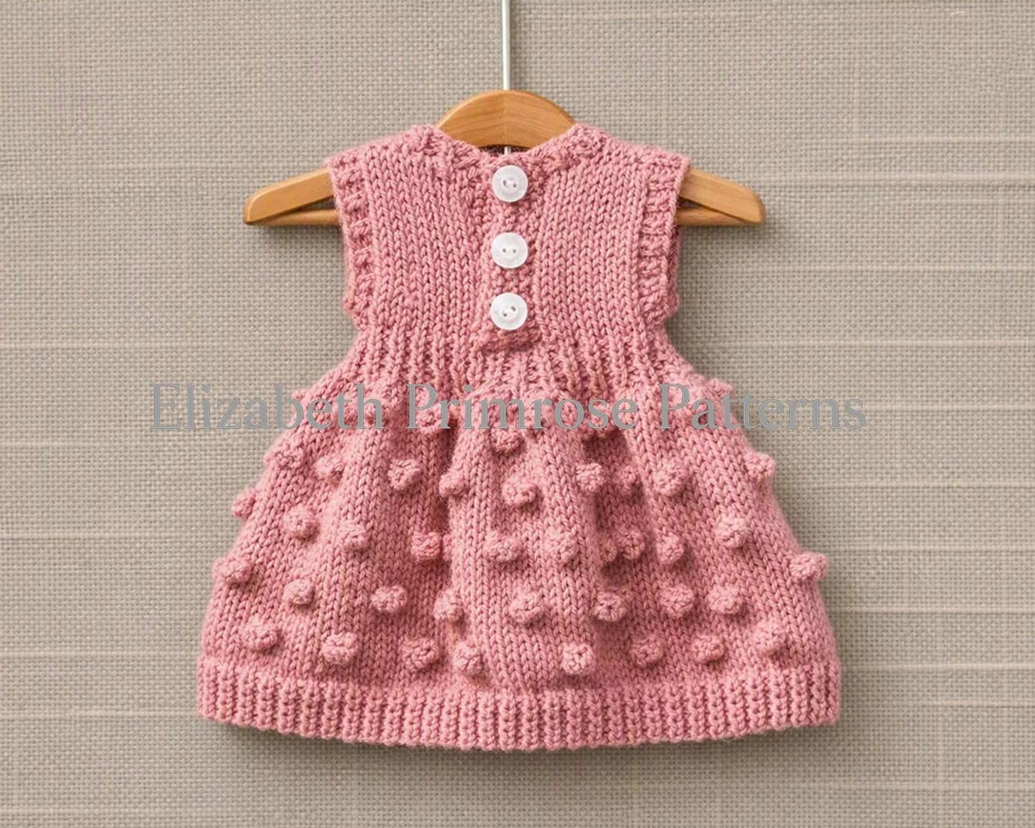Back view of a sleeveless pink knitted doll dress featuring three white buttons down the bodice and a textured bobble stitch skirt on a wooden hanger.