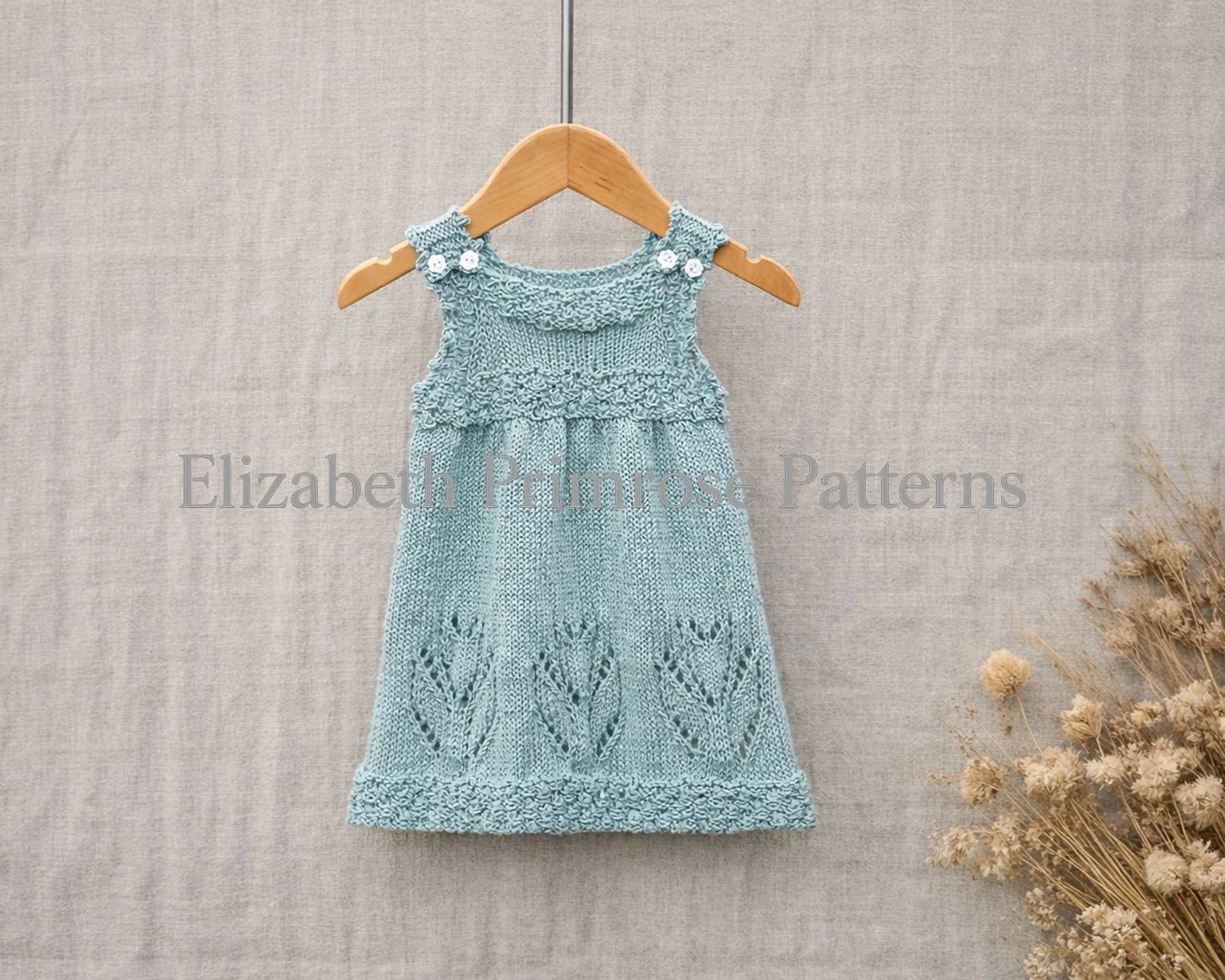 Light blue knitted baby dress on a wooden hanger with crisp lace tulip motifs and bobble details against a neutral fabric background.
