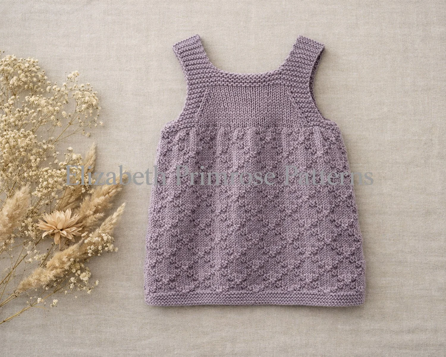 Textured lavender baby pinafore dress with buttoned straps displayed on a beige linen flatlay with muted dried flowers. Diamond lattice design. Elizabeth Primrose Patterns knitting pattern.