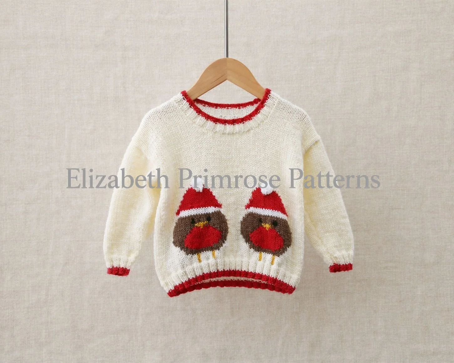 Cream children’s knitted jumper with two robins in Santa hats, photographed on a wooden hanger against a soft beige fabric background with festive red trims.